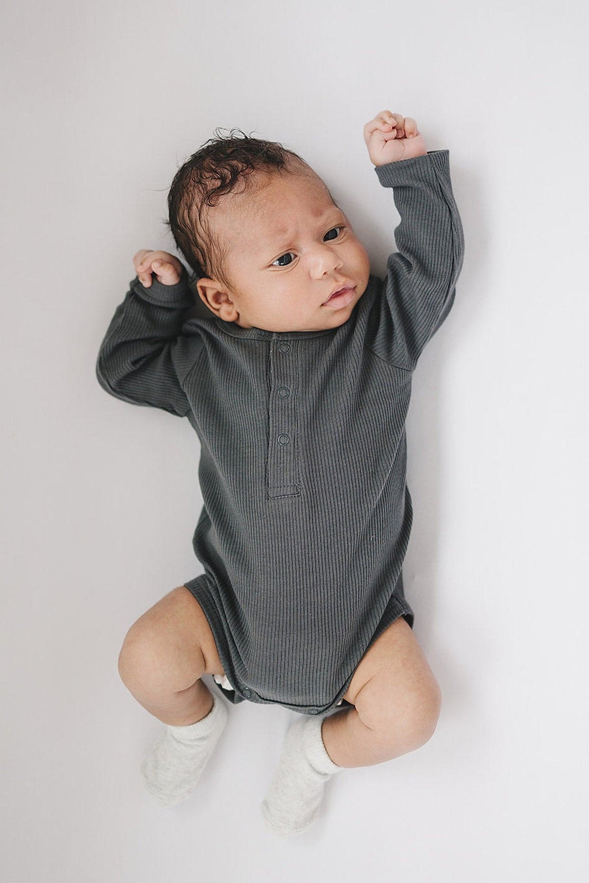 Baby wearing a charcoal ribbed long sleeve bodysuit with snaps, lying on a white surface, wearing white socks.