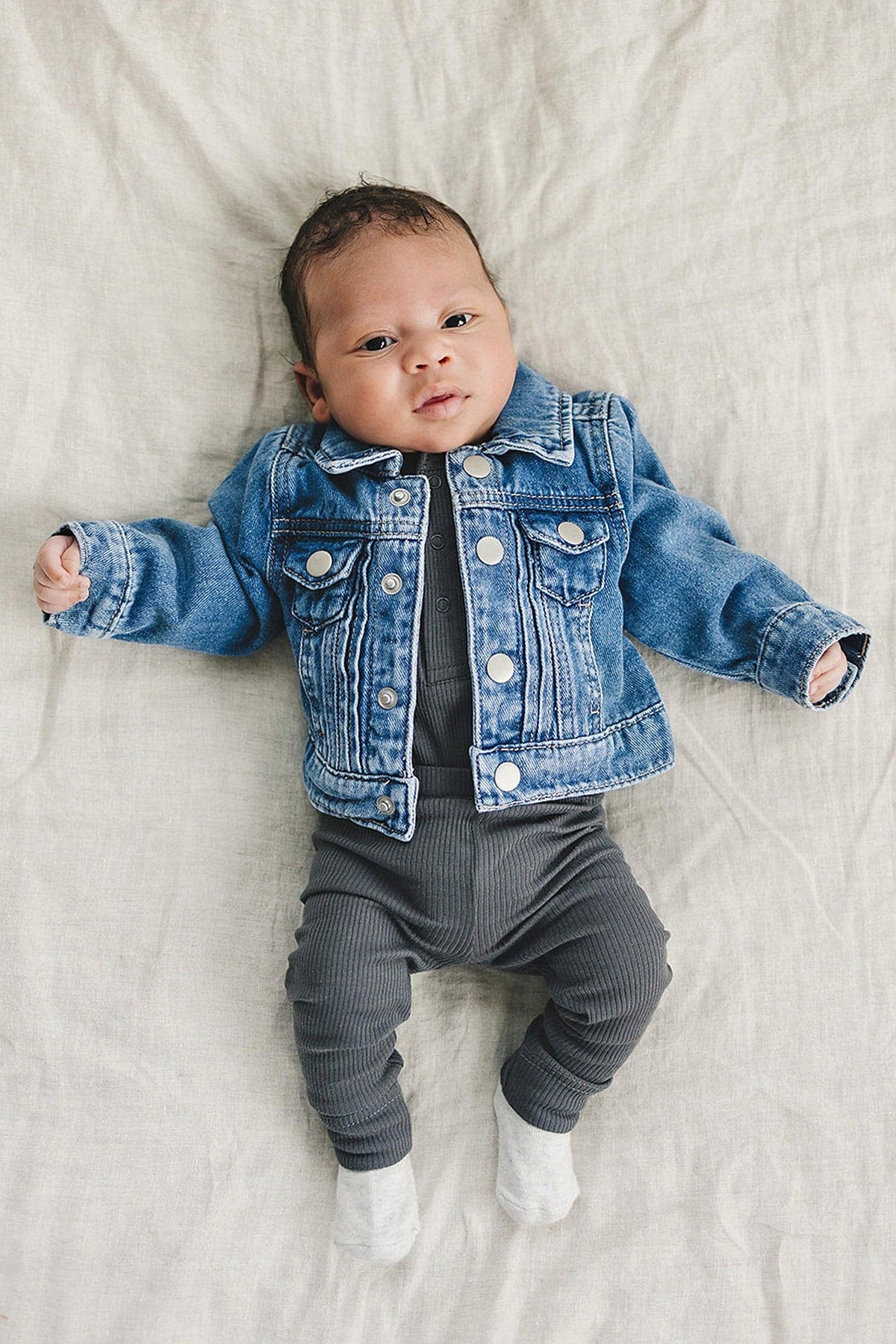 A baby wearing a denim jacket over a charcoal ribbed bodysuit, lying on a beige fabric surface.