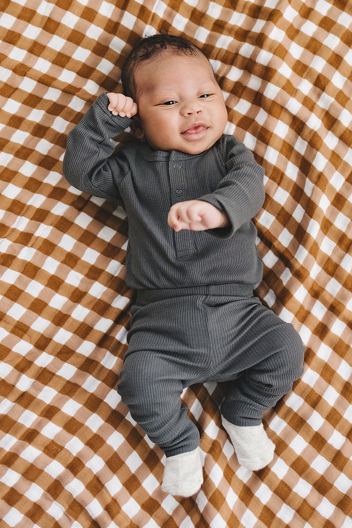 A baby wearing a charcoal ribbed long sleeve bodysuit lies on a brown and white checkered blanket.