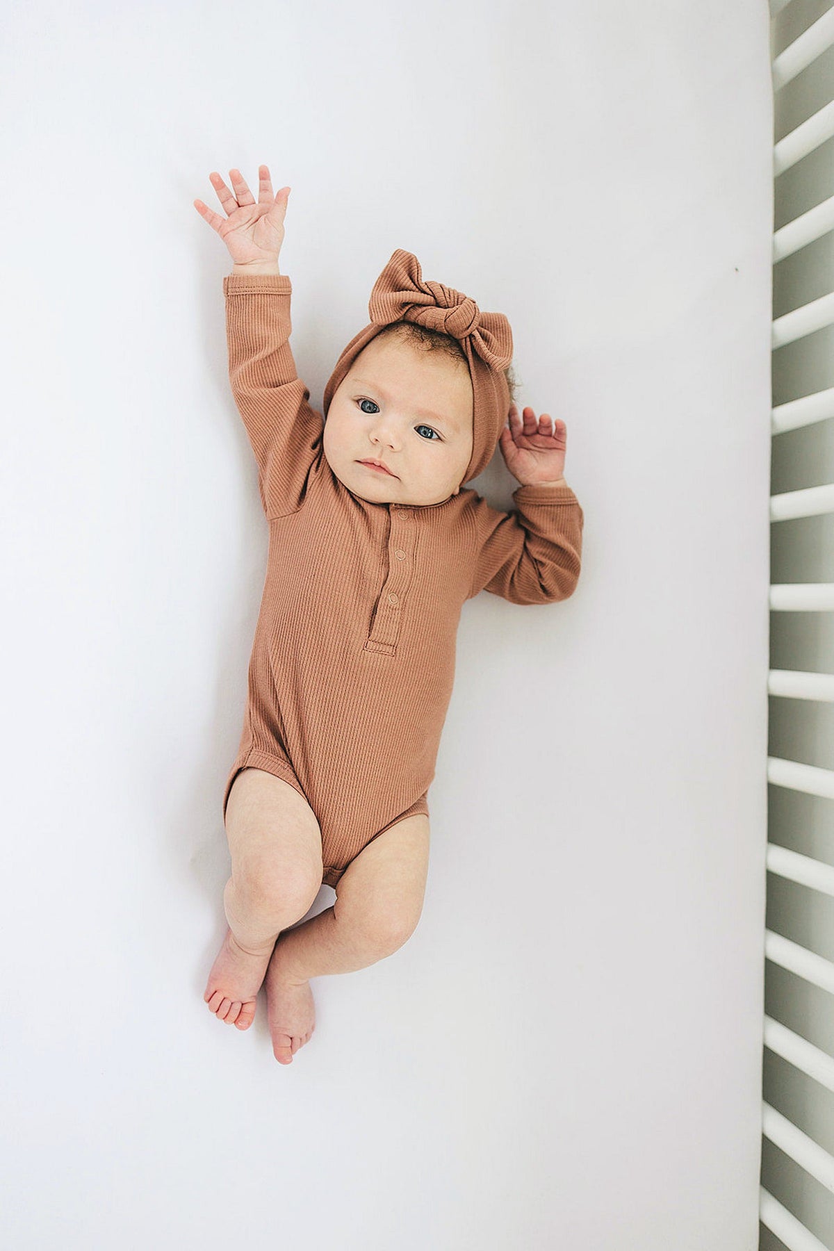 Baby in a dusty rose ribbed bodysuit with long sleeves and matching headband, lying in a crib.