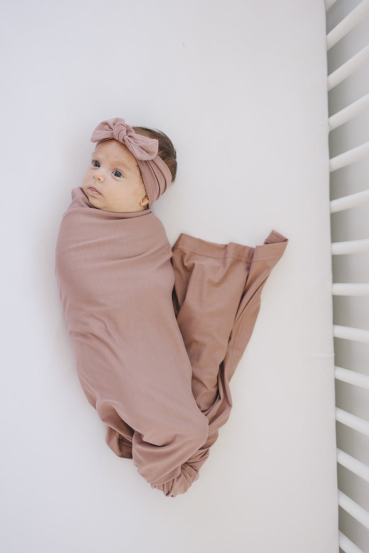 A baby wrapped in a dusty rose bamboo swaddle with a matching head wrap, lying in a crib.