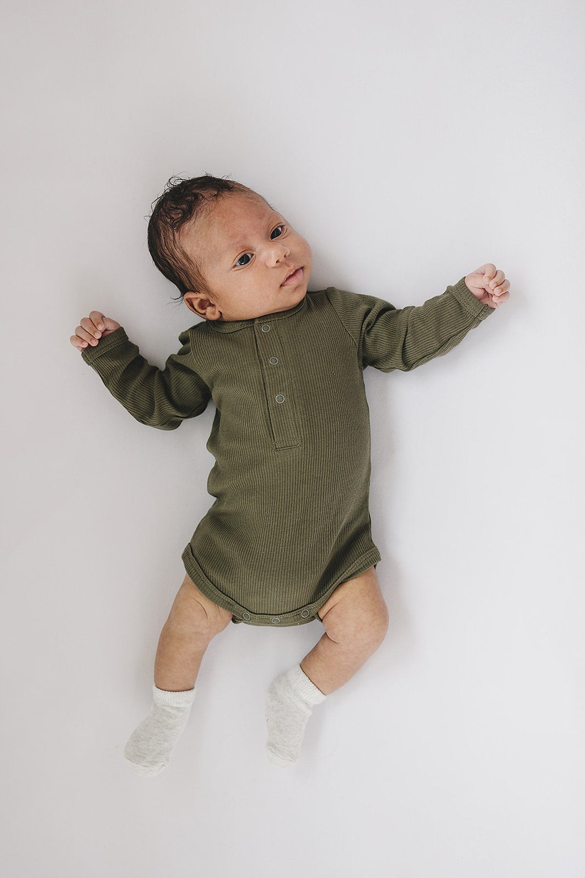 A baby wearing a green ribbed long sleeve bodysuit with snaps, lying on a white surface.