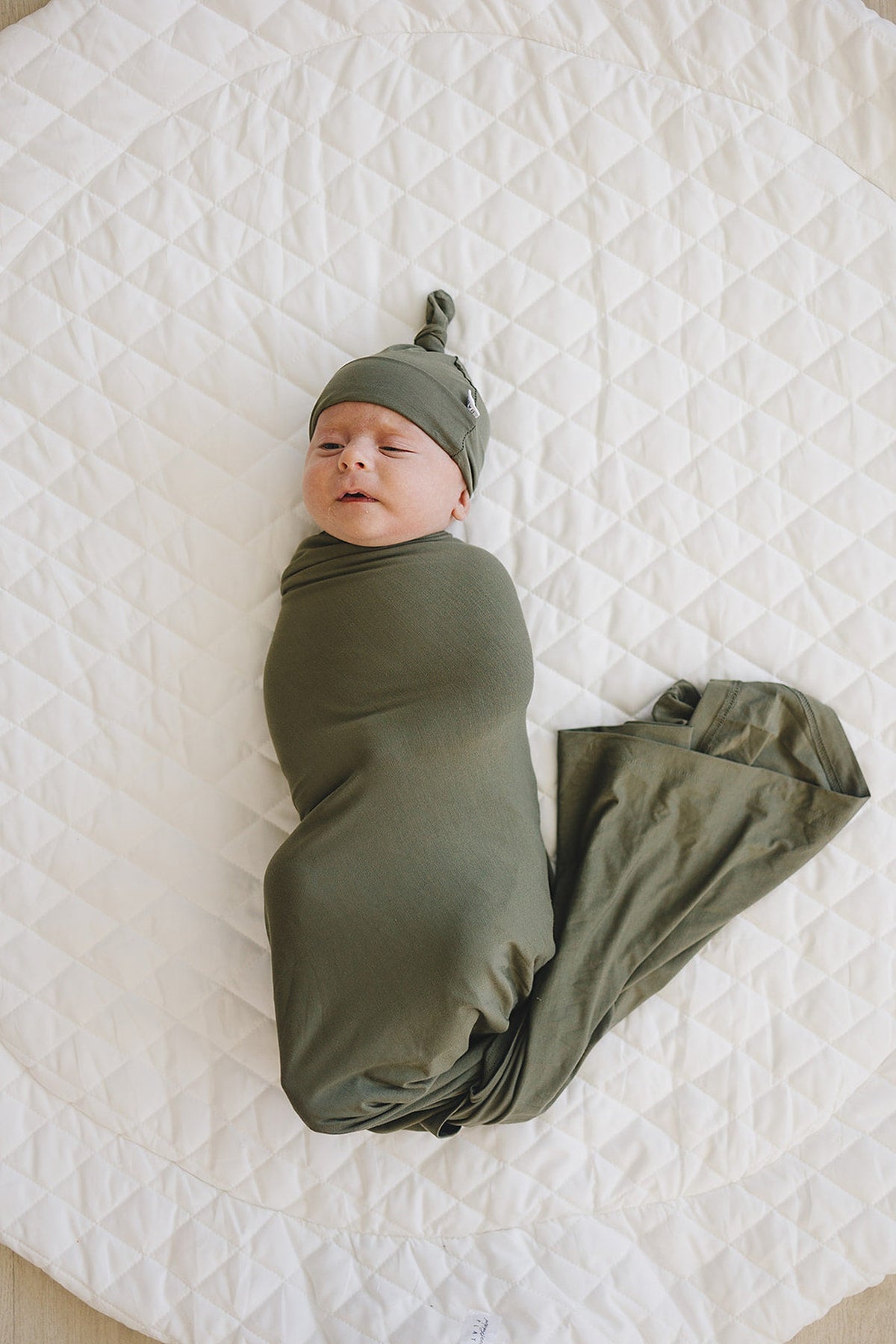 Newborn wrapped in an olive green swaddle and hat, lying on a quilted white blanket.