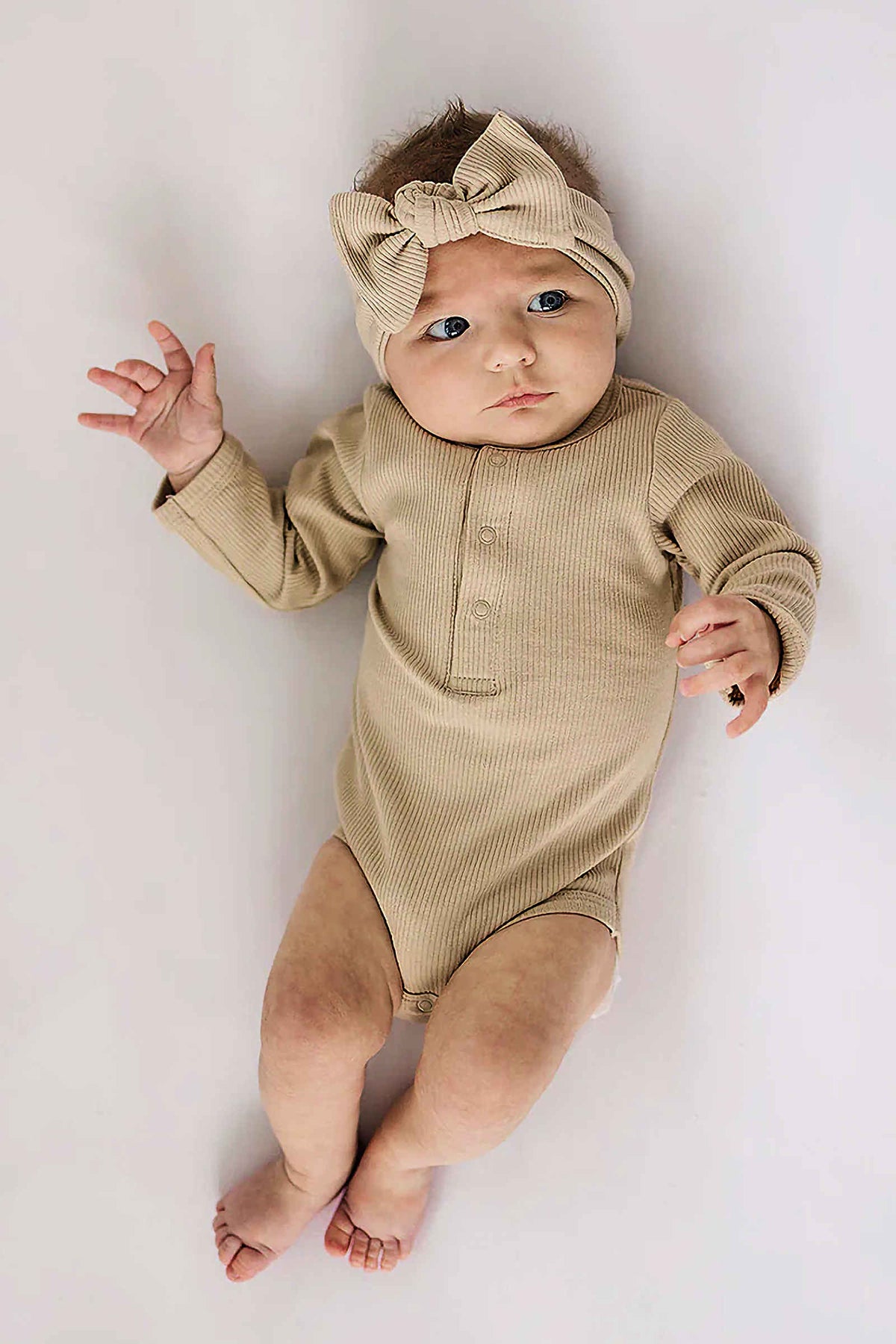 A baby wearing a ribbed oatmeal-colored long sleeve bodysuit with snaps and a matching bow headband lies on a white.