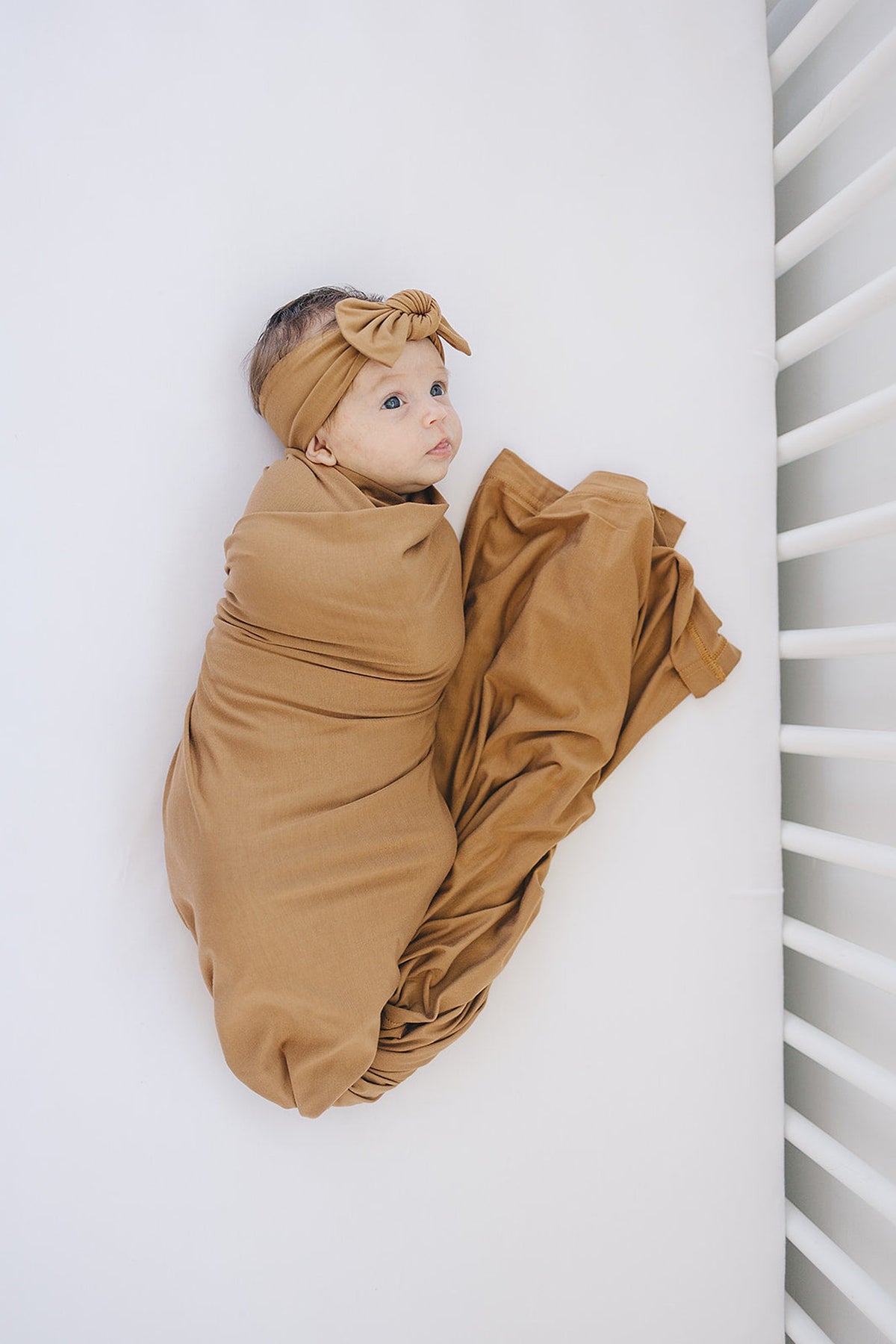 A baby is wrapped in a mustard-colored bamboo fabric swaddle with a matching head wrap, lying in a crib.