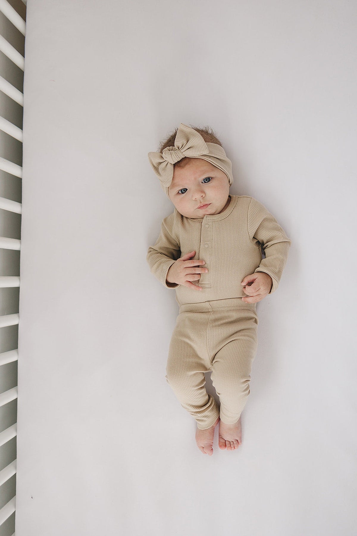 A baby wearing an oatmeal-colored ribbed bodysuit and matching headband lies on a white crib mattress.
