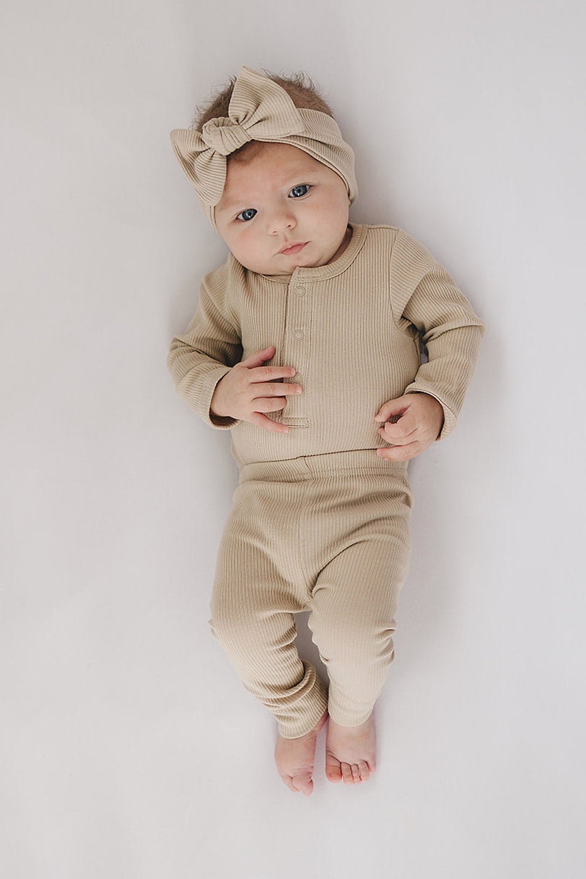 Baby wearing oatmeal-colored ribbed outfit and matching headband, lying on white surface, looking upward.