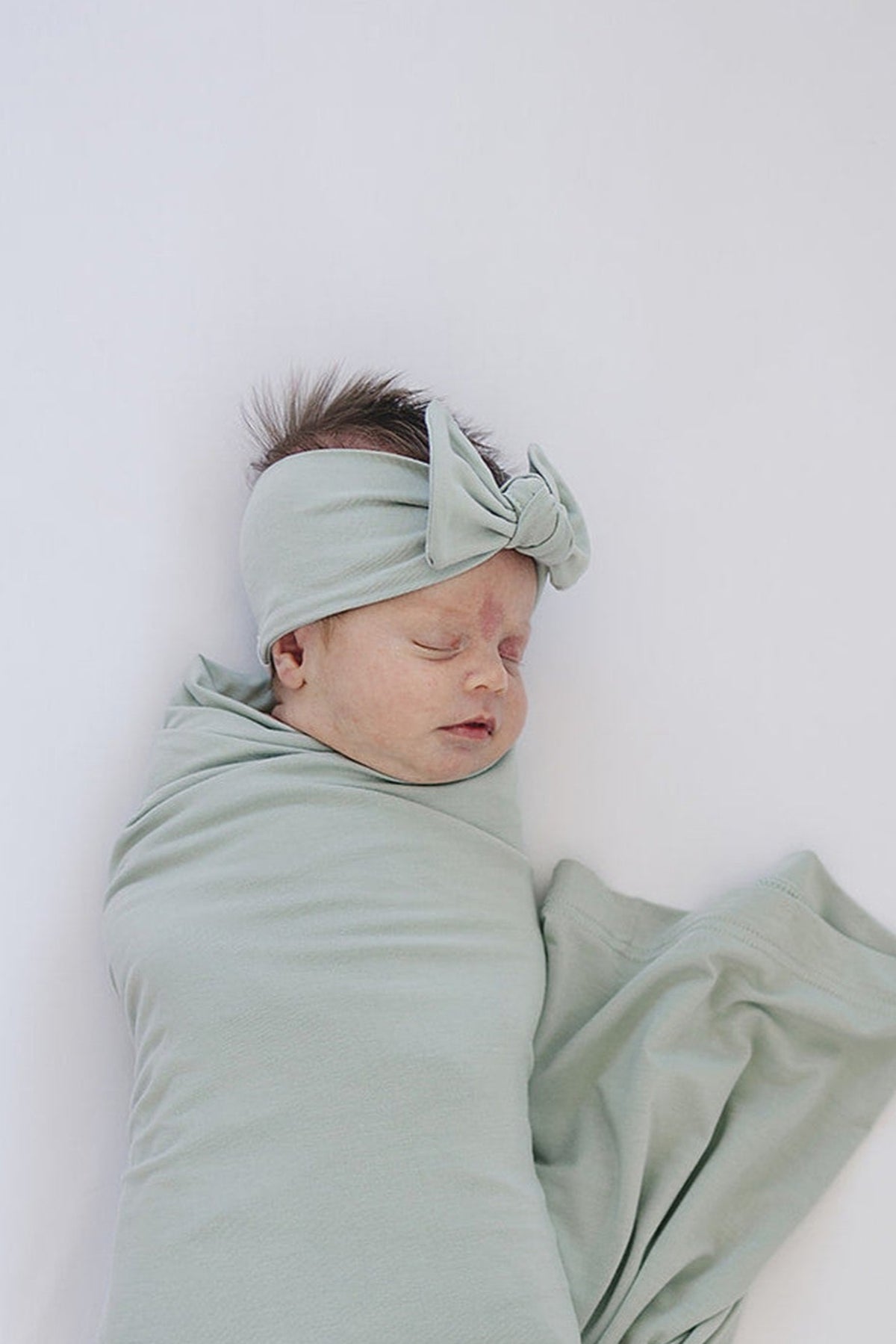 A baby wrapped in a soft sage bamboo blanket, wearing a matching sage head wrap with a bow.