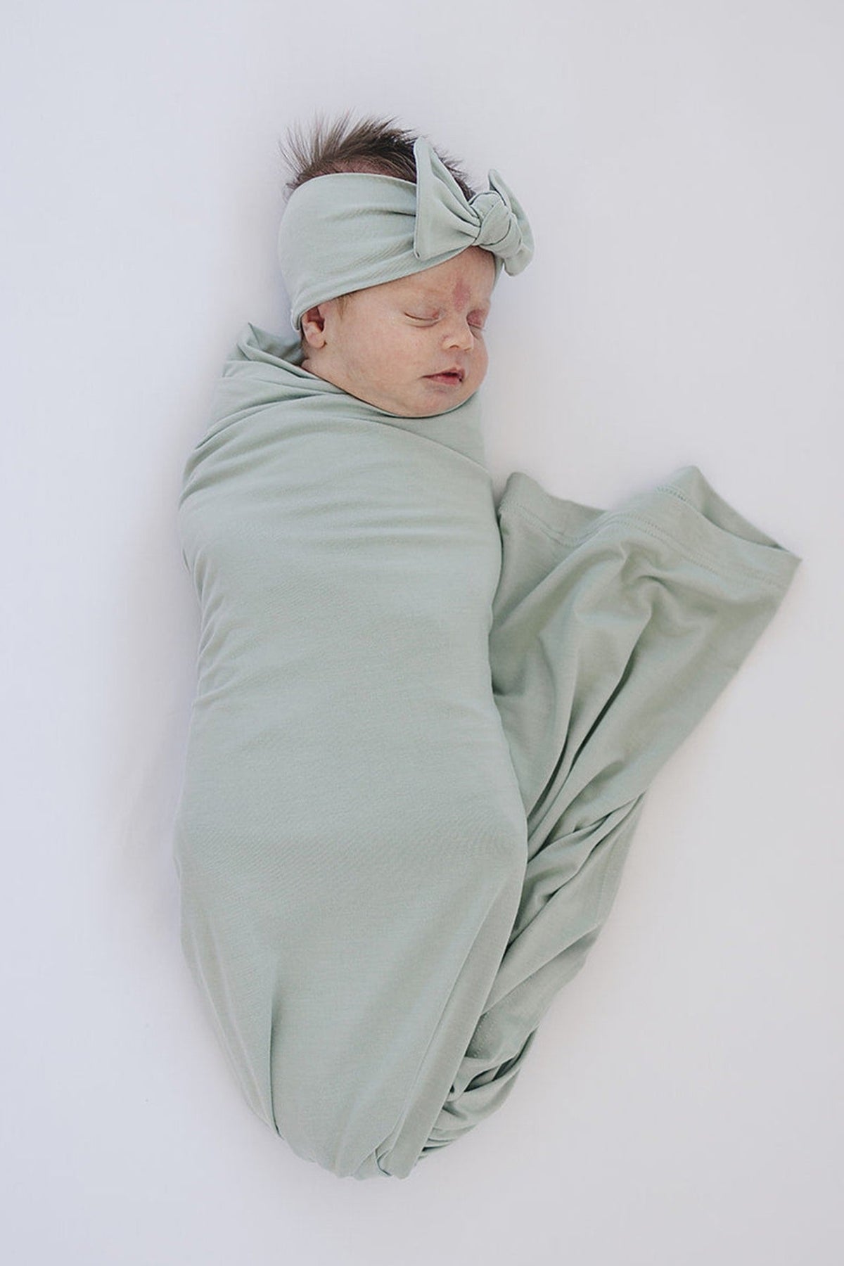 A baby is wrapped in a sage green swaddle blanket with a matching headband, lying on a white surface.