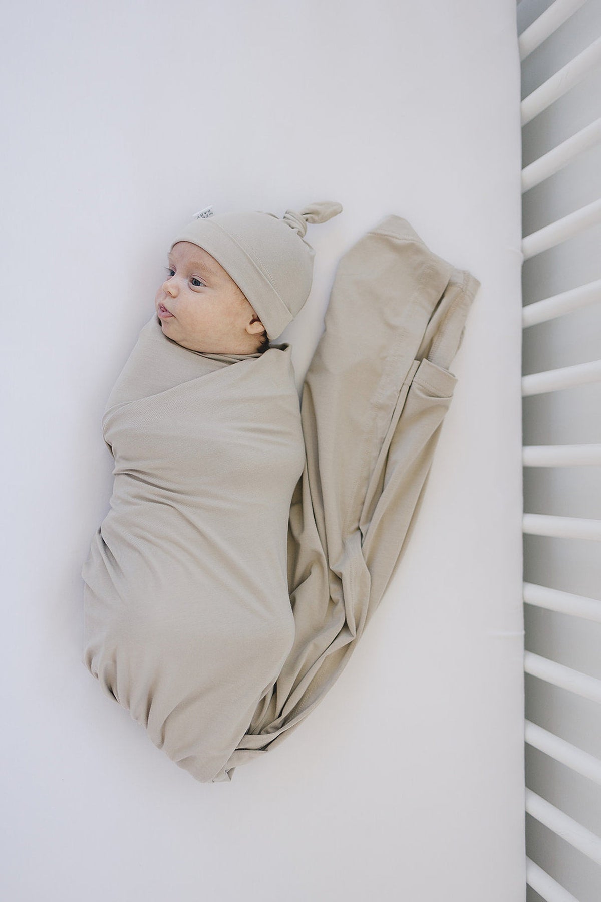 A baby wrapped in a beige bamboo swaddle with a matching knotted hat, lying on a white surface.
