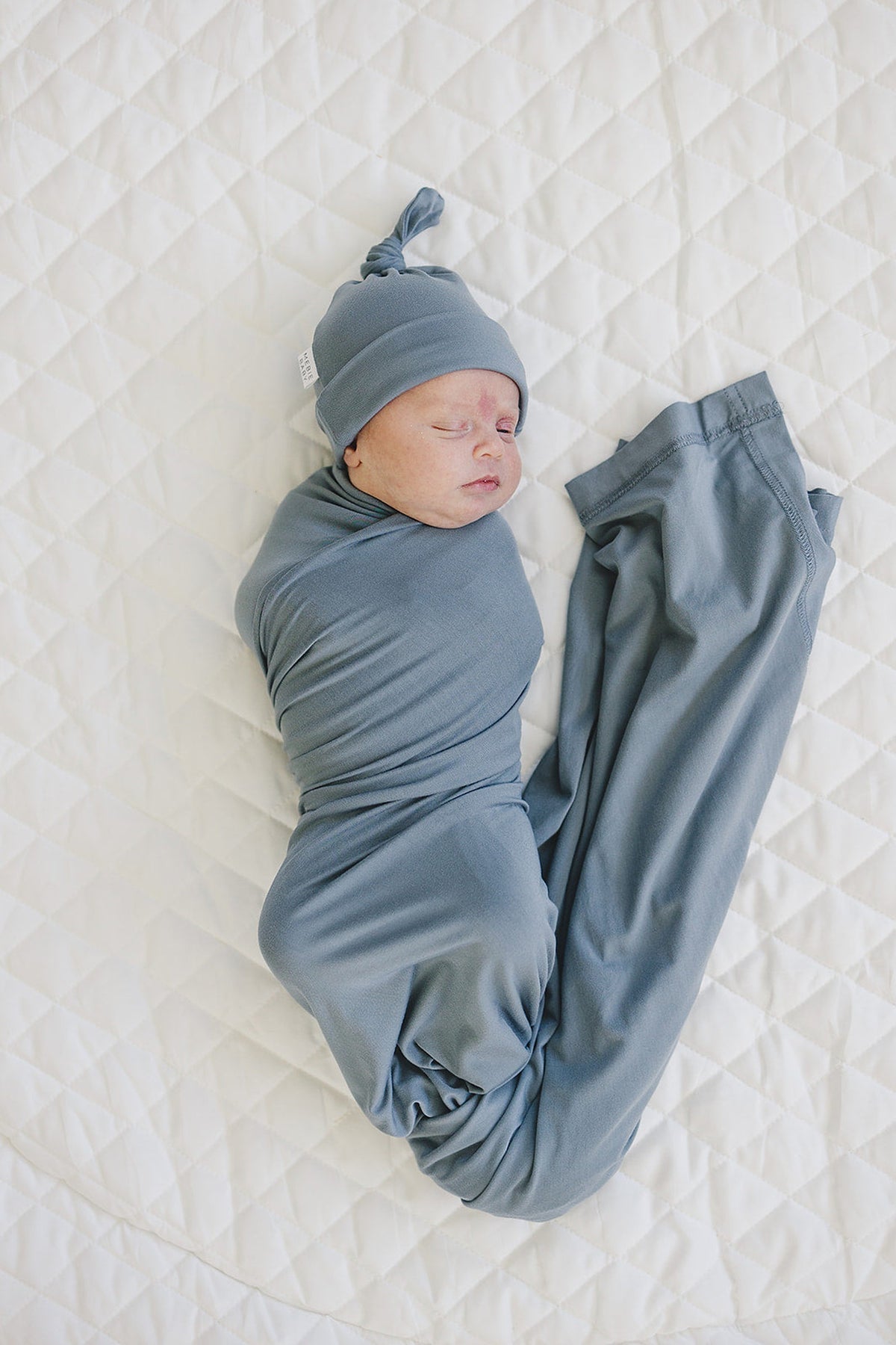 A newborn baby is wrapped in a dusty blue swaddle and knot hat, lying on a quilted white blanket.