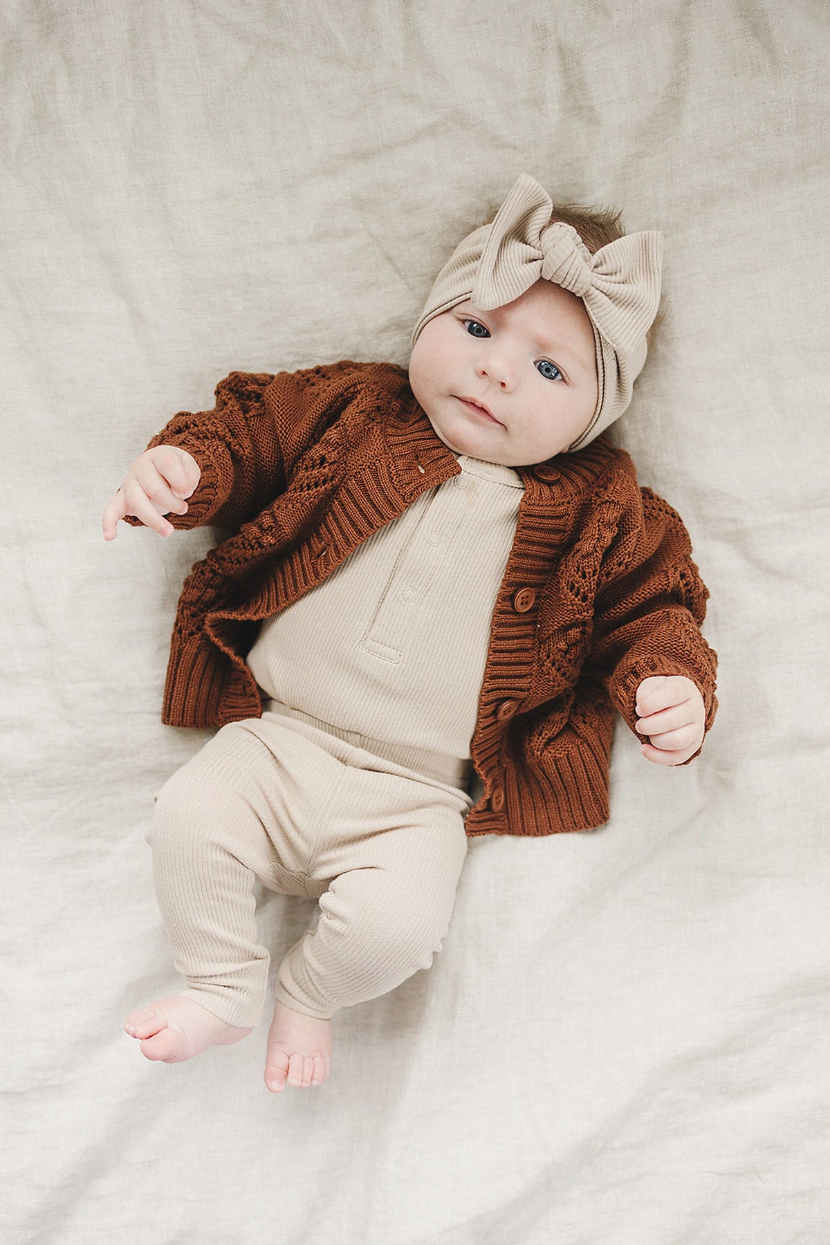 A baby is wearing a ribbed beige bodysuit with a matching headband and a brown knitted cardigan.
