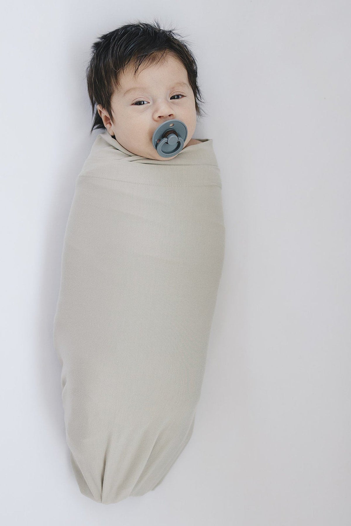 A baby with dark hair is wrapped in a beige swaddle blanket, using a gray pacifier.