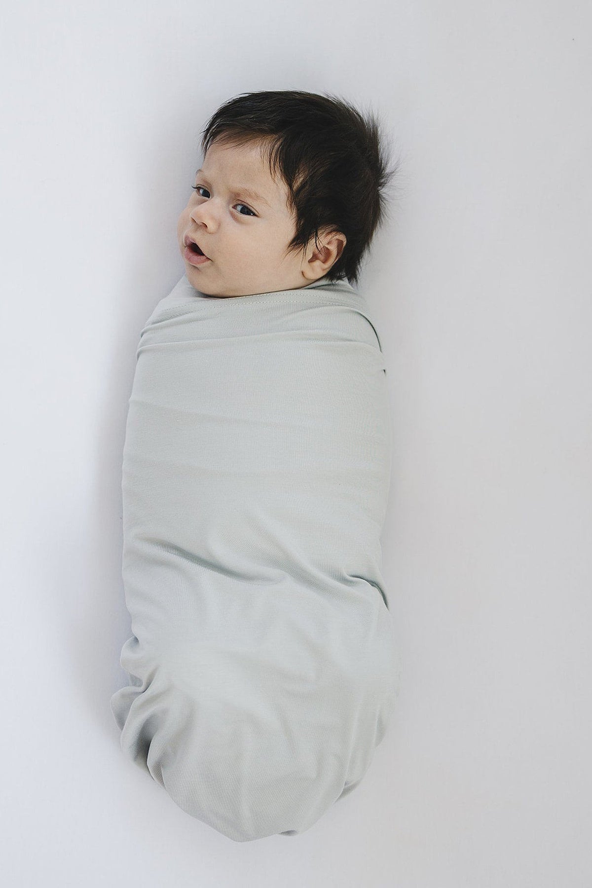 A baby is swaddled in a light grey bamboo blanket, lying on a white surface.