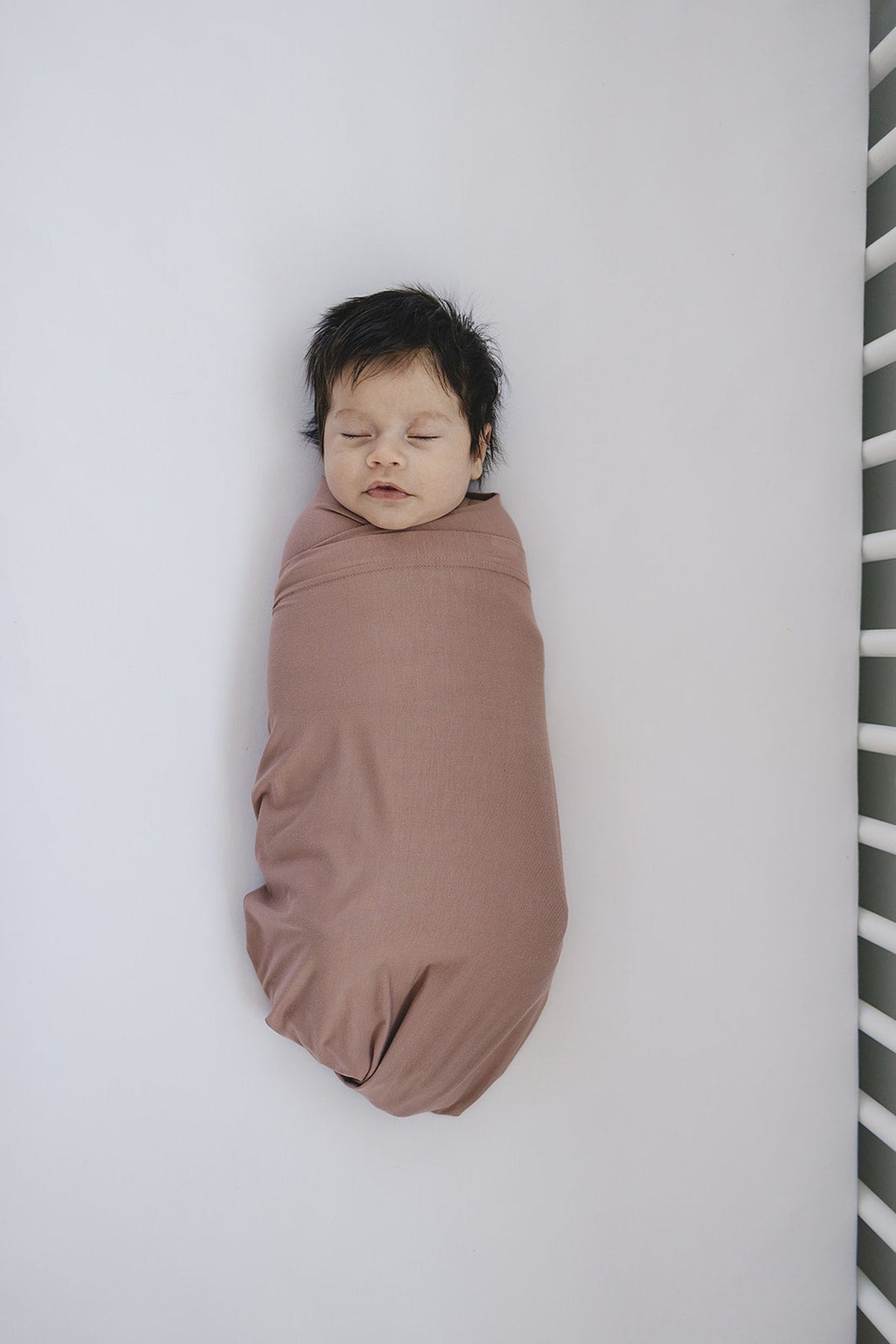 A baby sleeps peacefully wrapped in a dusty rose swaddle blanket, lying on a white crib mattress.