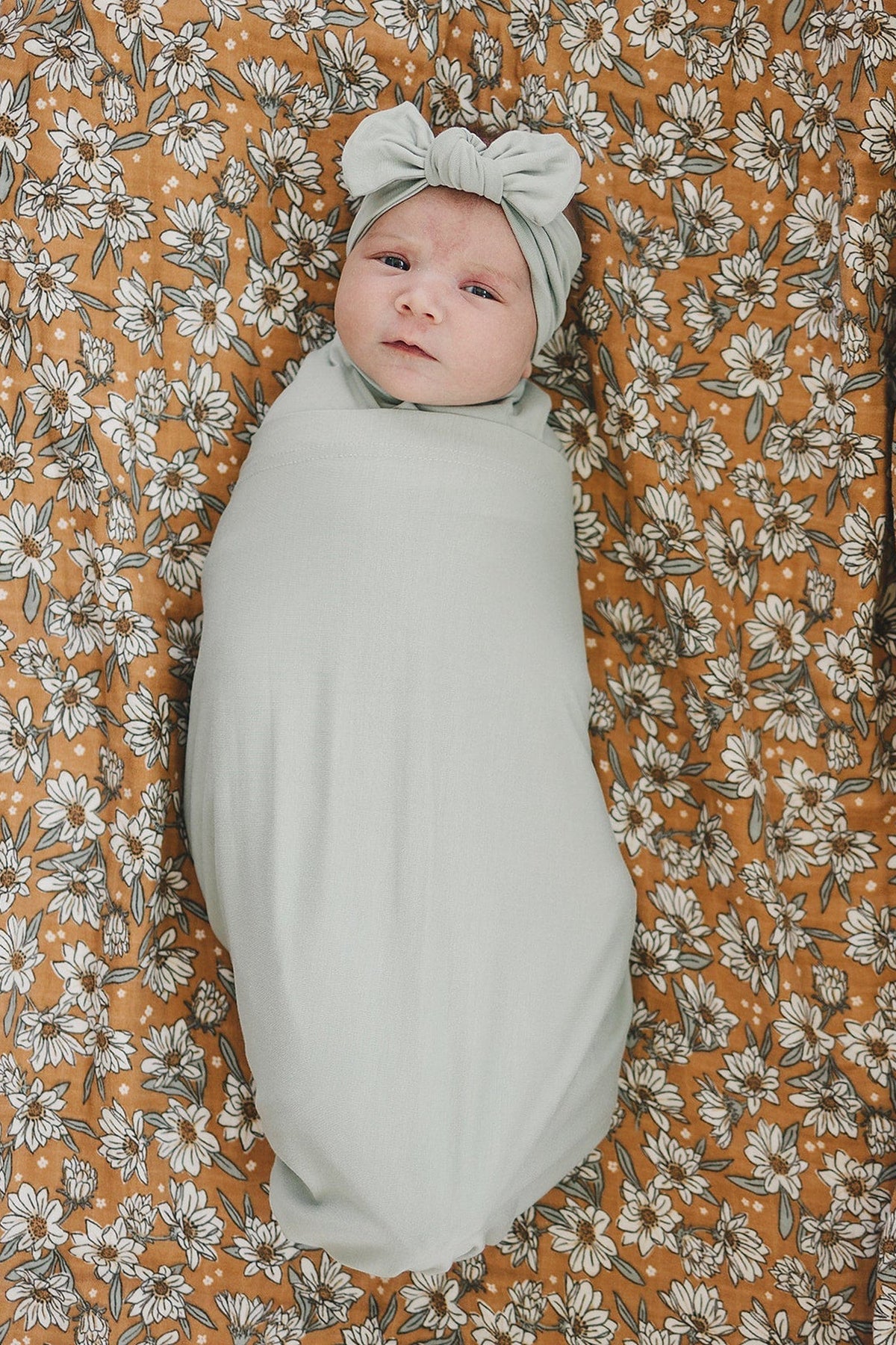 A baby in a sage green swaddle and matching headband, lying on a floral-patterned blanket.