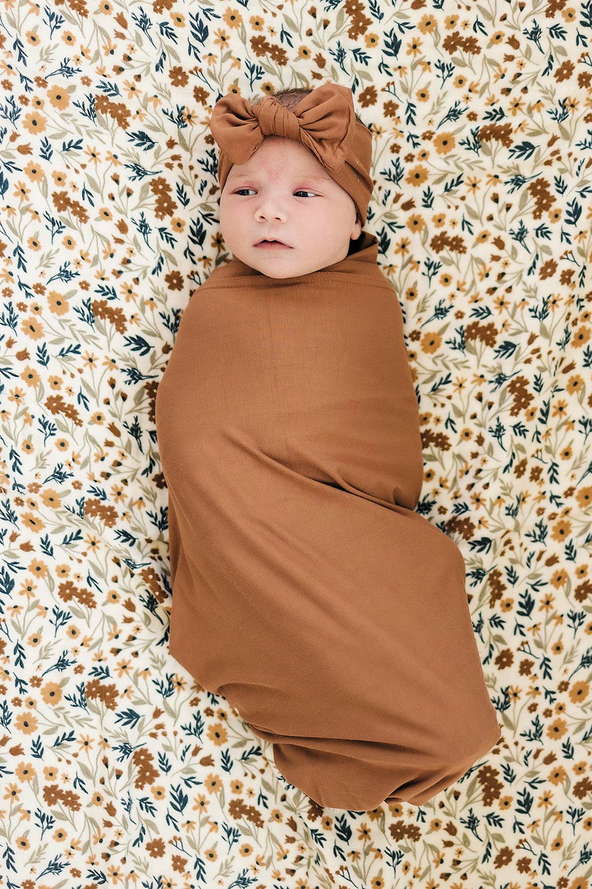 A baby in a rust-colored bamboo swaddle and matching hat lies on a floral-patterned backdrop.