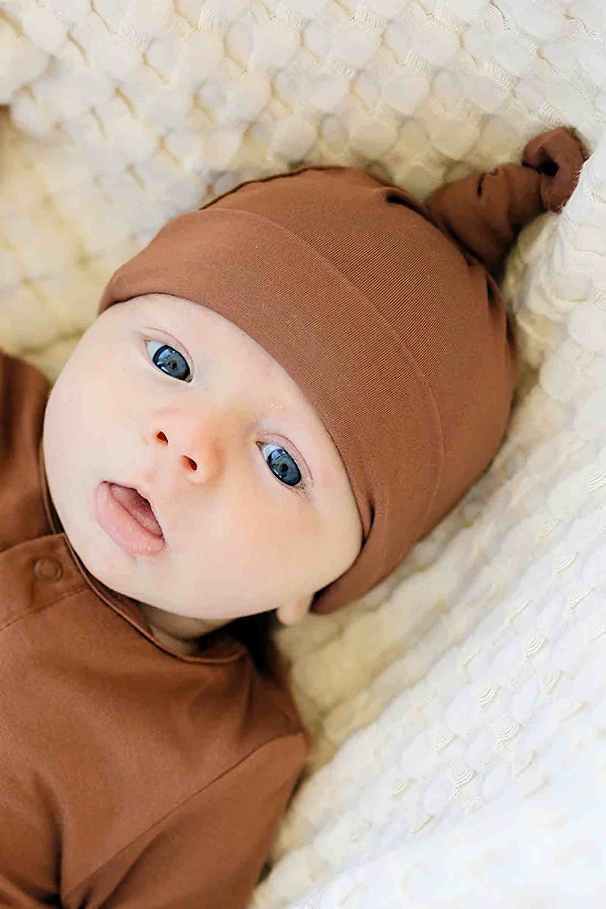 A baby wearing a rust-colored hat and outfit lies on a textured white blanket, gazing upwards.