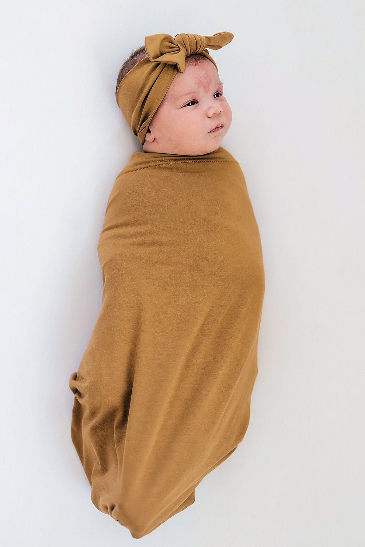 Newborn in a mustard bamboo swaddle with matching head wrap, lying on a white background.