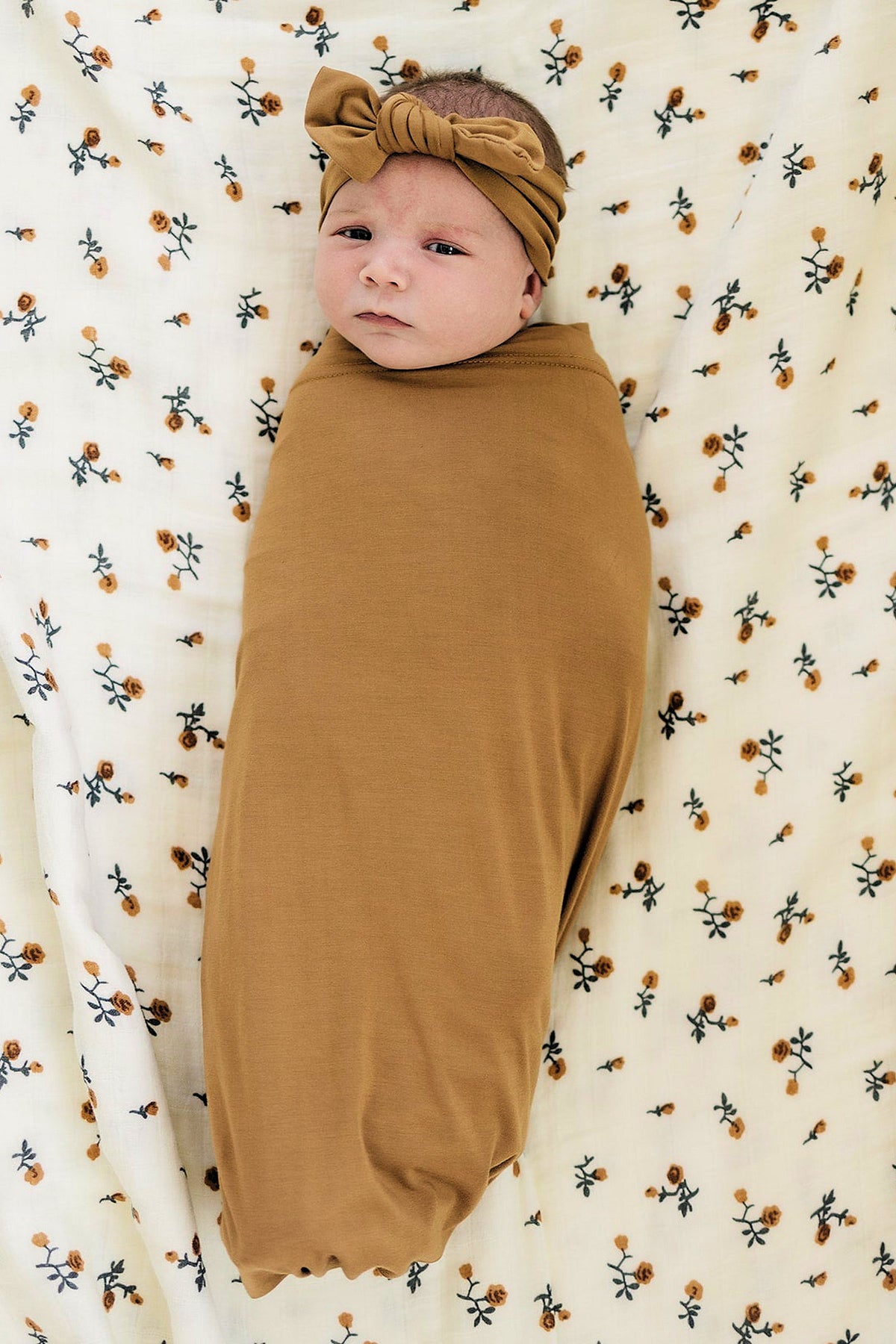 A newborn wrapped in a mustard swaddle and matching headband, lying on a floral-patterned blanket.