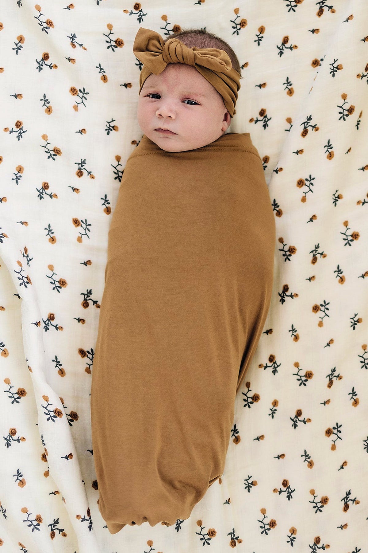 A baby wrapped in a mustard-colored swaddle with matching headband, lying on a floral-patterned blanket.