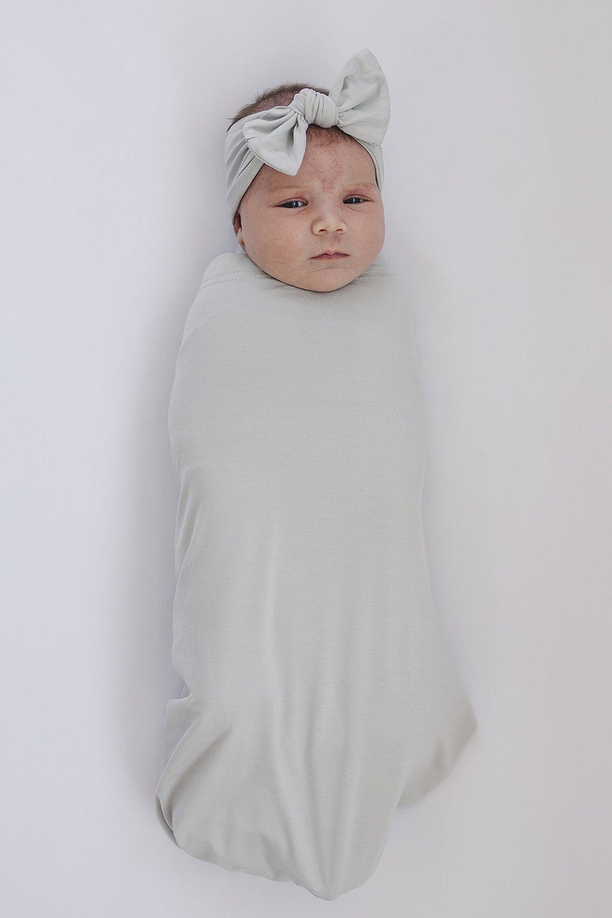 A baby is swaddled in a soft, light gray fabric with a matching bow head wrap on a white background.