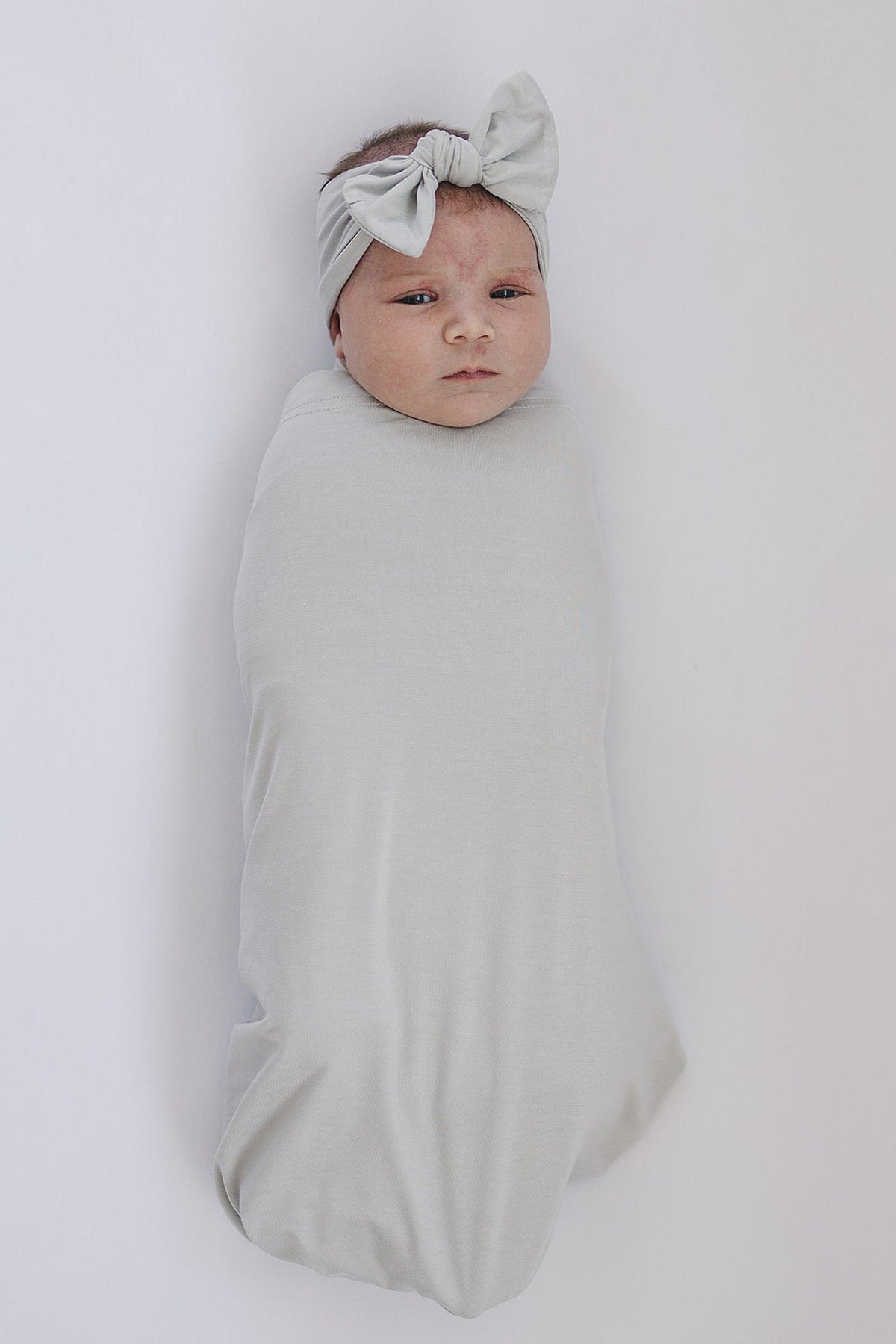 A baby wrapped in a light gray swaddle blanket with a matching bow headband, lying on a white surface.