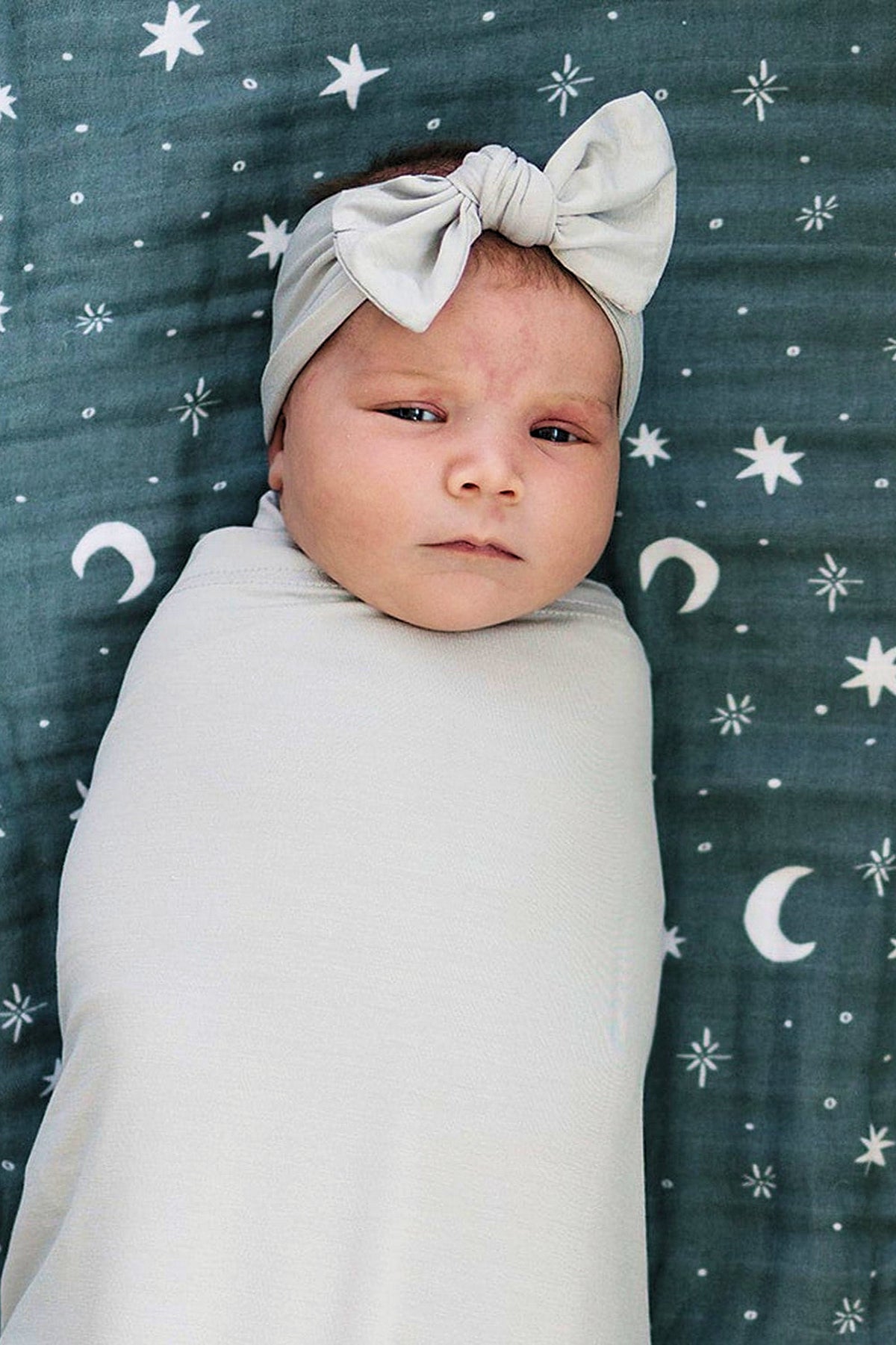 A baby wrapped in a light fabric, wearing a head wrap with a bow, on a starry patterned blanket.