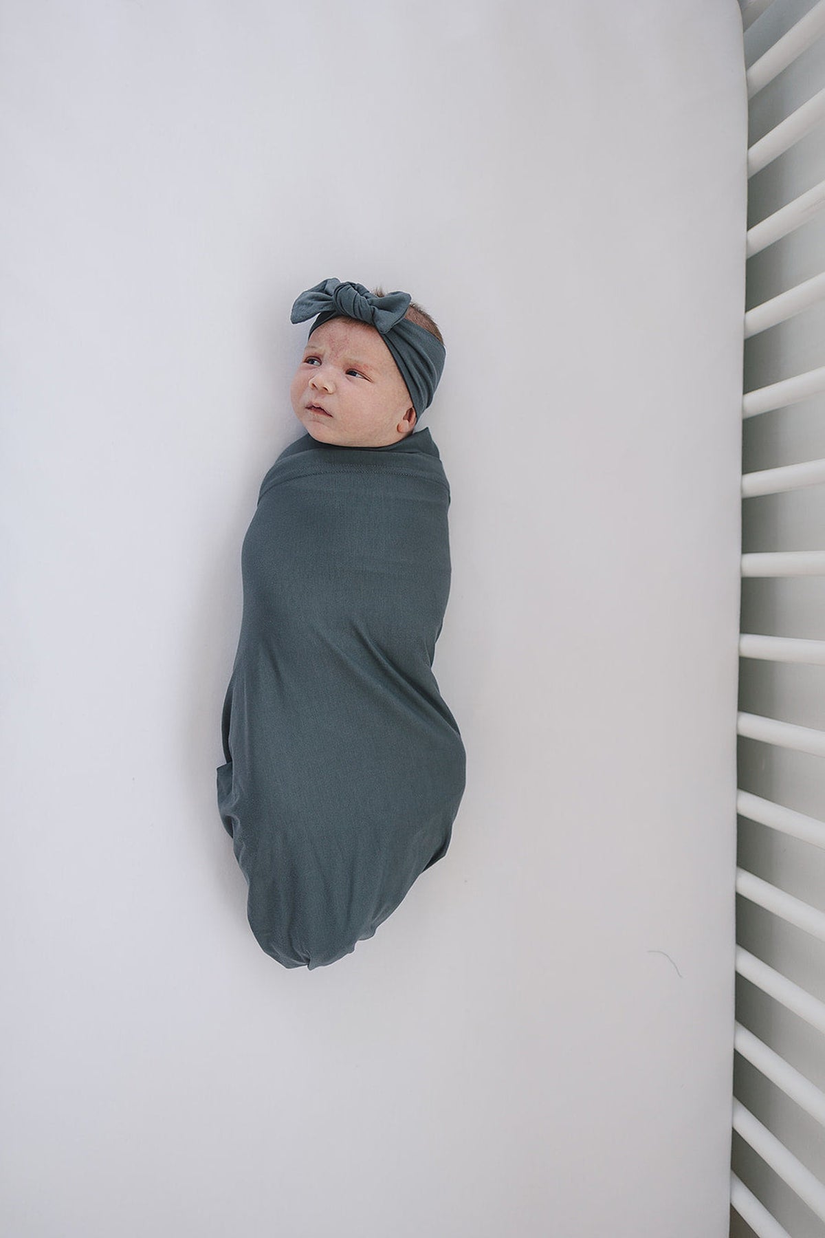 Newborn baby wrapped in a charcoal swaddle with a matching head wrap, lying in a white crib.