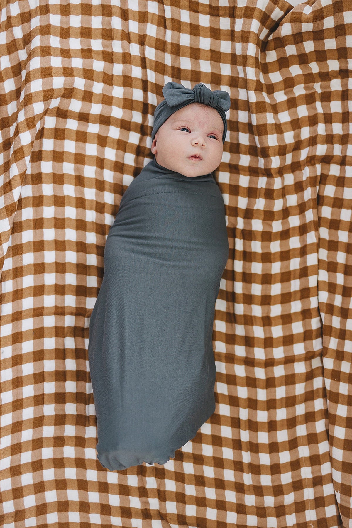 A baby wrapped in a charcoal stretch swaddle blanket lies on a brown and white checkered fabric.