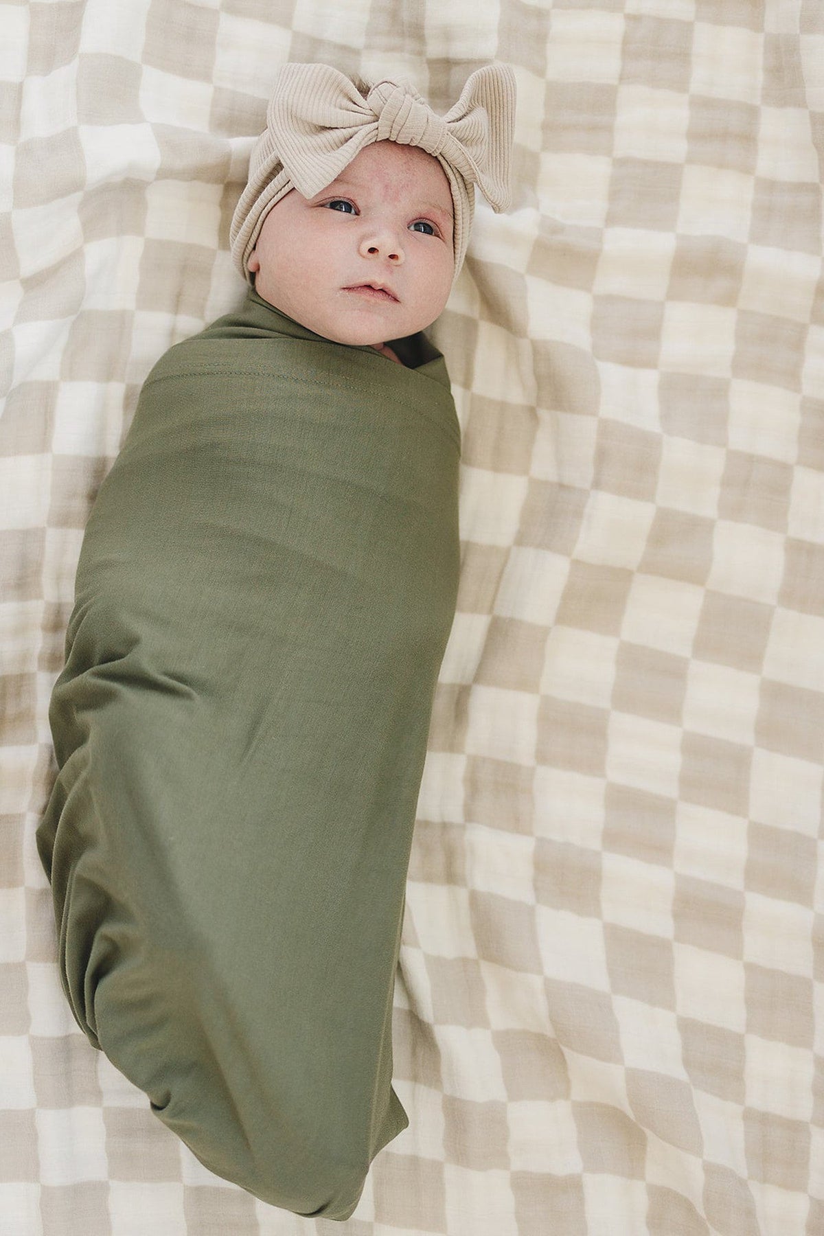 A baby wrapped in an olive green swaddle lies on a checkered blanket, wearing a beige bow headband.