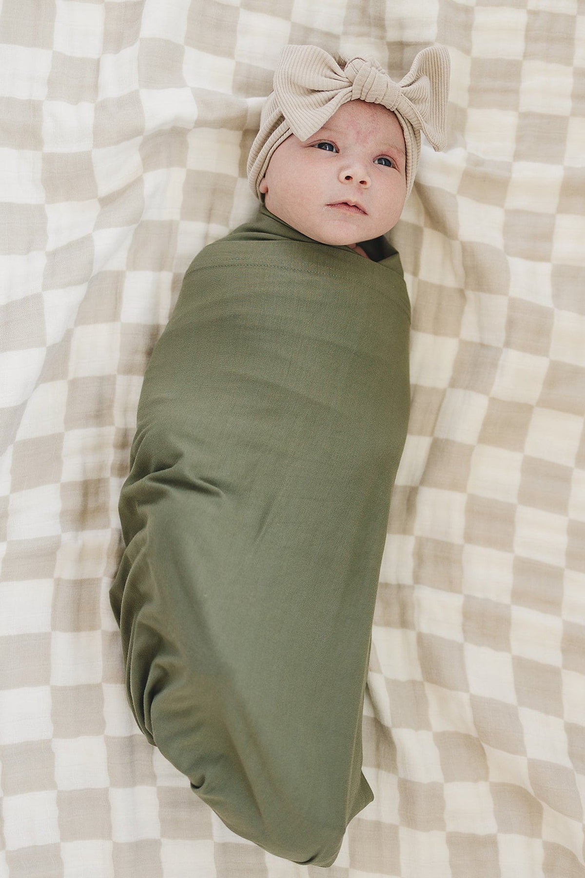 A baby in a green swaddle lies on a taupe checkered muslin quilt, wearing a beige bow headband.