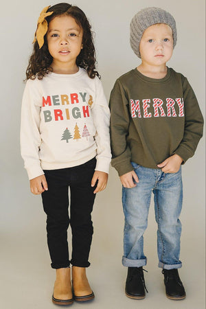 A girl in a cream sweater with MERRY & BRIGHT stands beside a boy in an olive green sweatshirt saying.