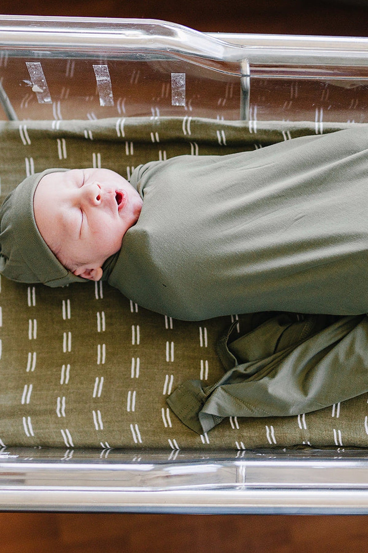 Newborn wrapped in an olive green swaddle blanket, lying in a hospital bassinet with a matching patterned sheet.