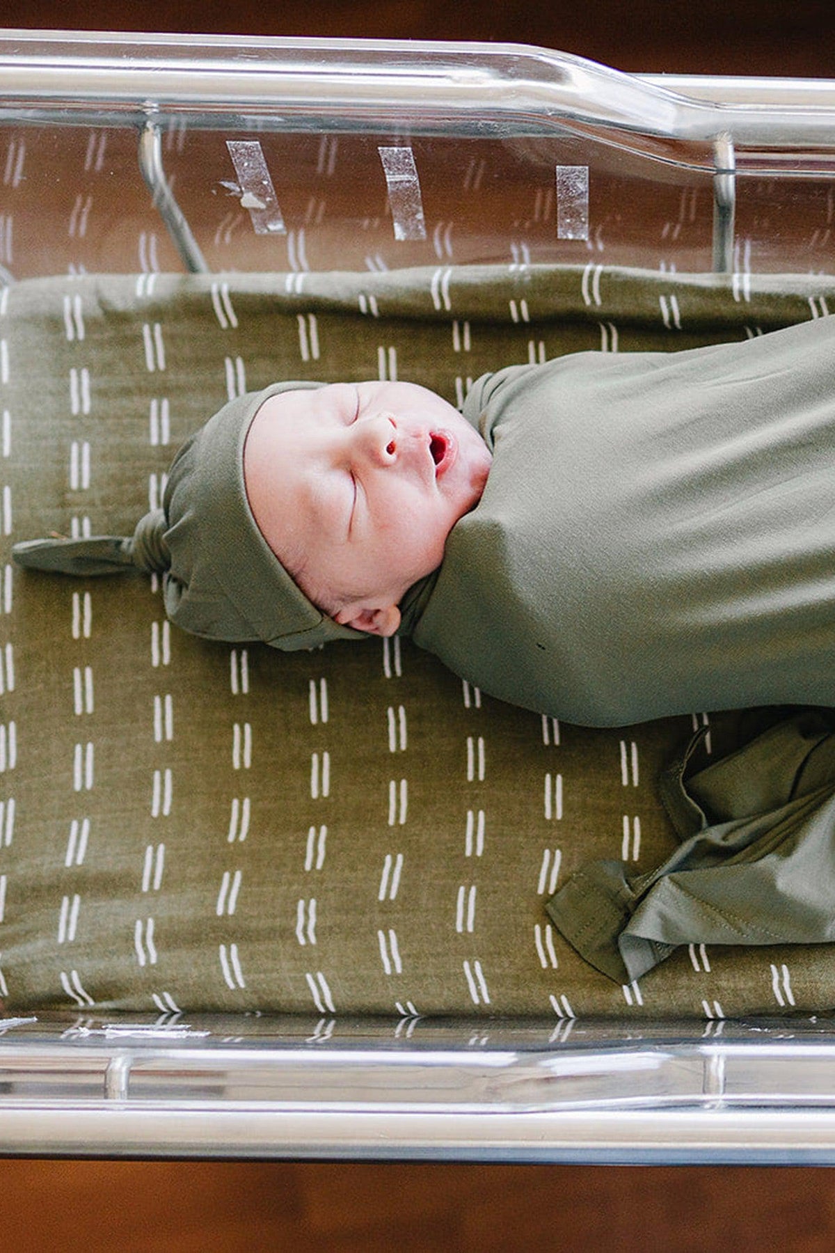 Newborn in olive bamboo swaddle and hat, lying in a crib with patterned bedding.