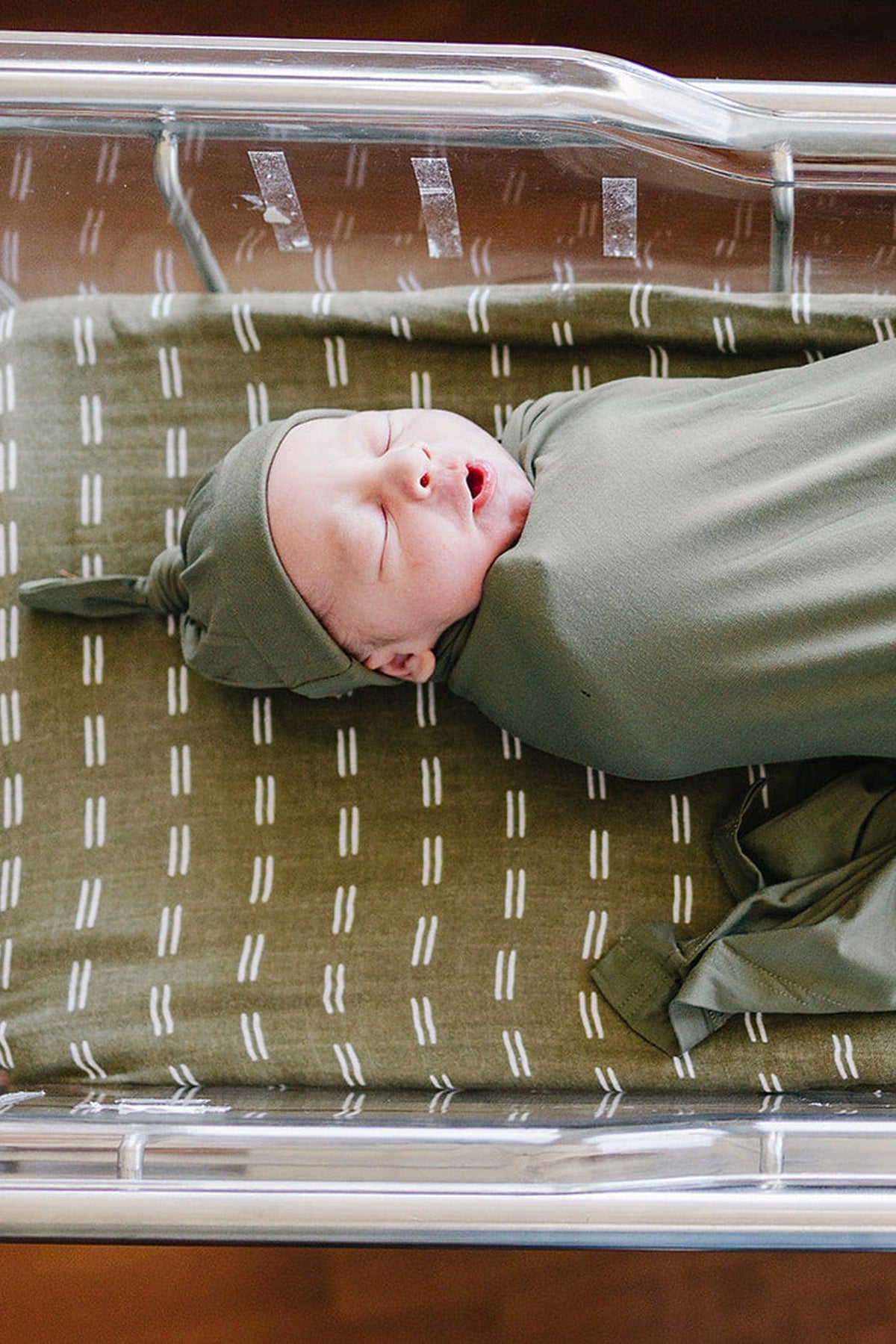 Newborn baby wearing an olive knot hat and swaddle, lying in a hospital bassinet on patterned bedding.