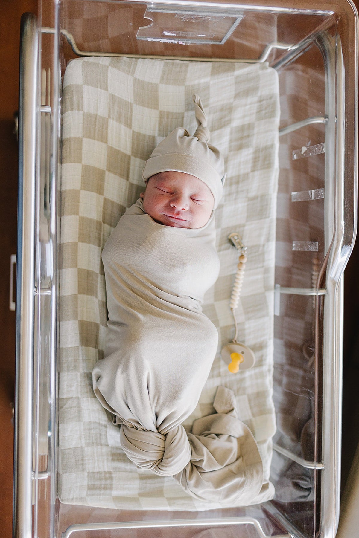 A newborn baby is wrapped in a beige swaddle and hat, lying in a hospital bassinet.