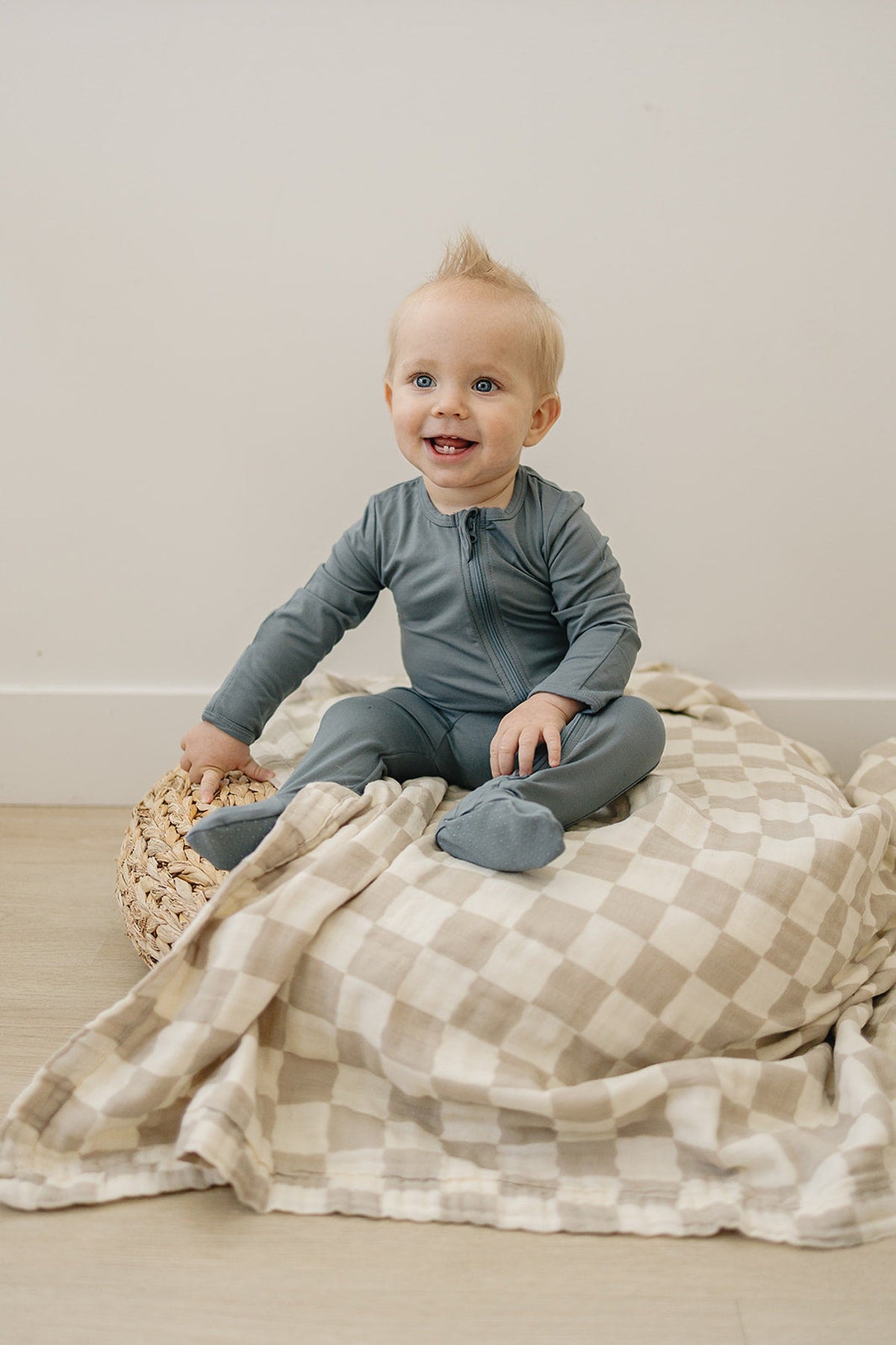 A baby in dusty blue footed pajamas sits on a checkered blanket, smiling happily.