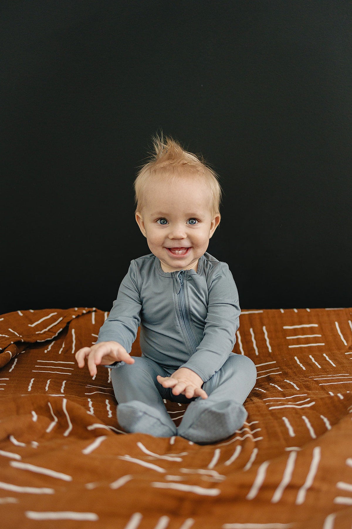 Smiling baby in dusty blue footed zipper pajamas sits on a patterned brown blanket with a black background.