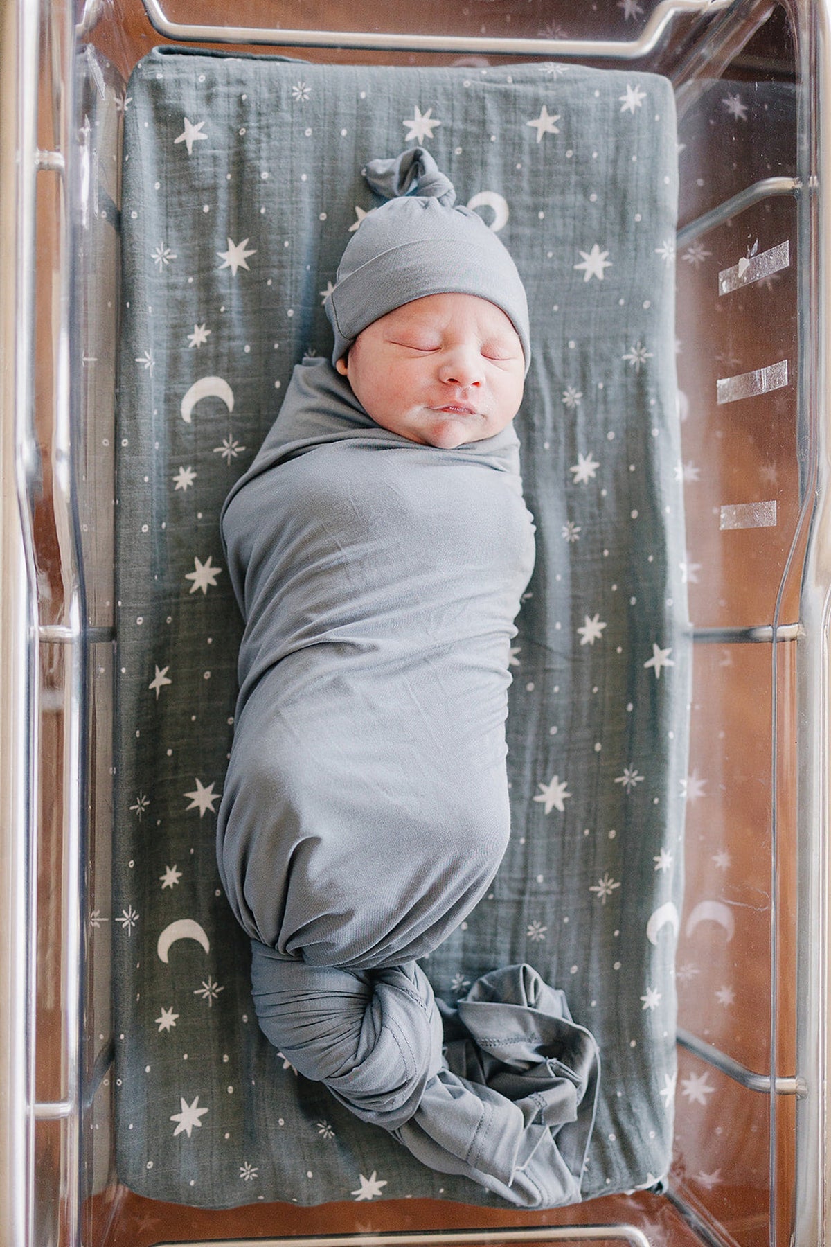 A newborn wrapped in a dusty blue bamboo swaddle with a matching hat, lying on a patterned blanket.
