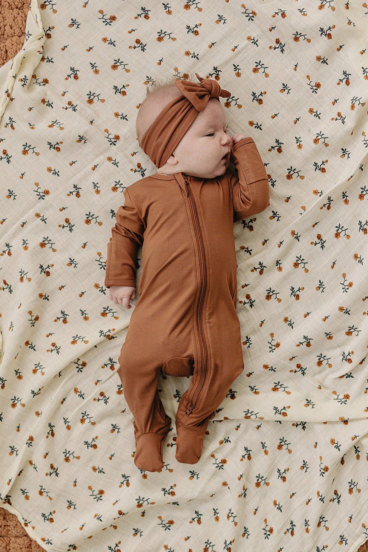 Baby in rust-colored footed pajama with zipper, lying on a floral-patterned blanket, wearing a matching headband.