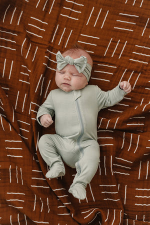 A baby in sage green footed pajamas and headband lies on a brown patterned blanket with white lines.