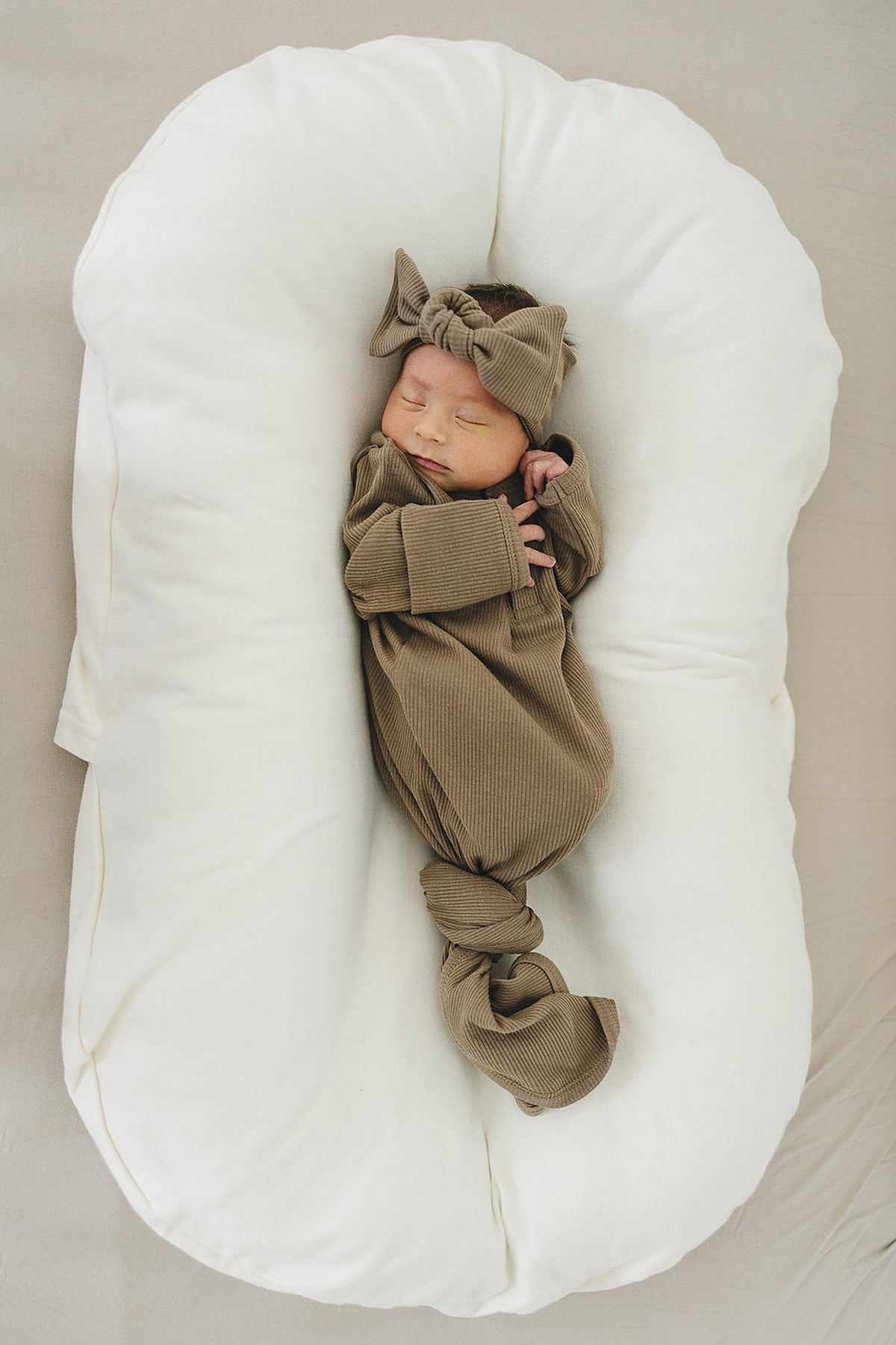 A baby sleeps in a white pillow wearing a cocoa ribbed outfit and matching head wrap with a bow.