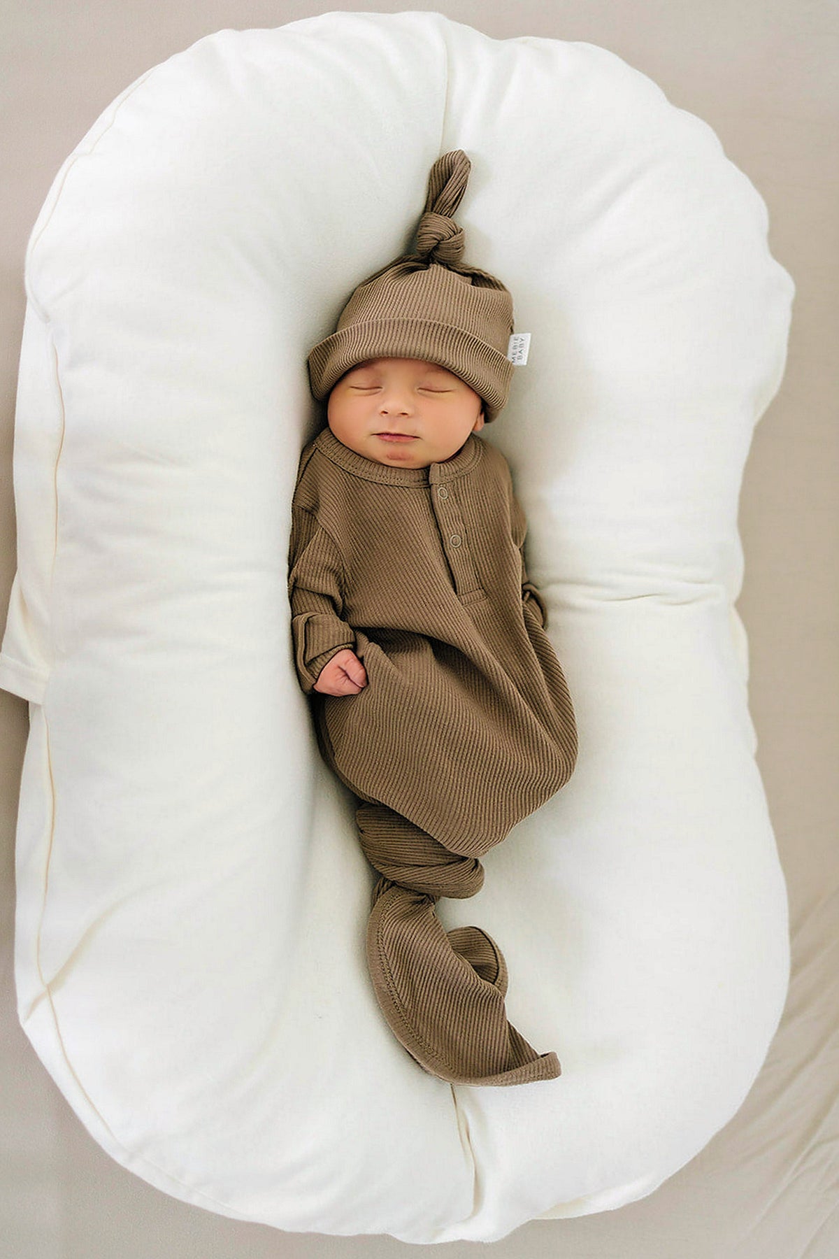 Newborn in a cocoa ribbed organic cotton gown and knot hat, resting on a soft white cushion.
