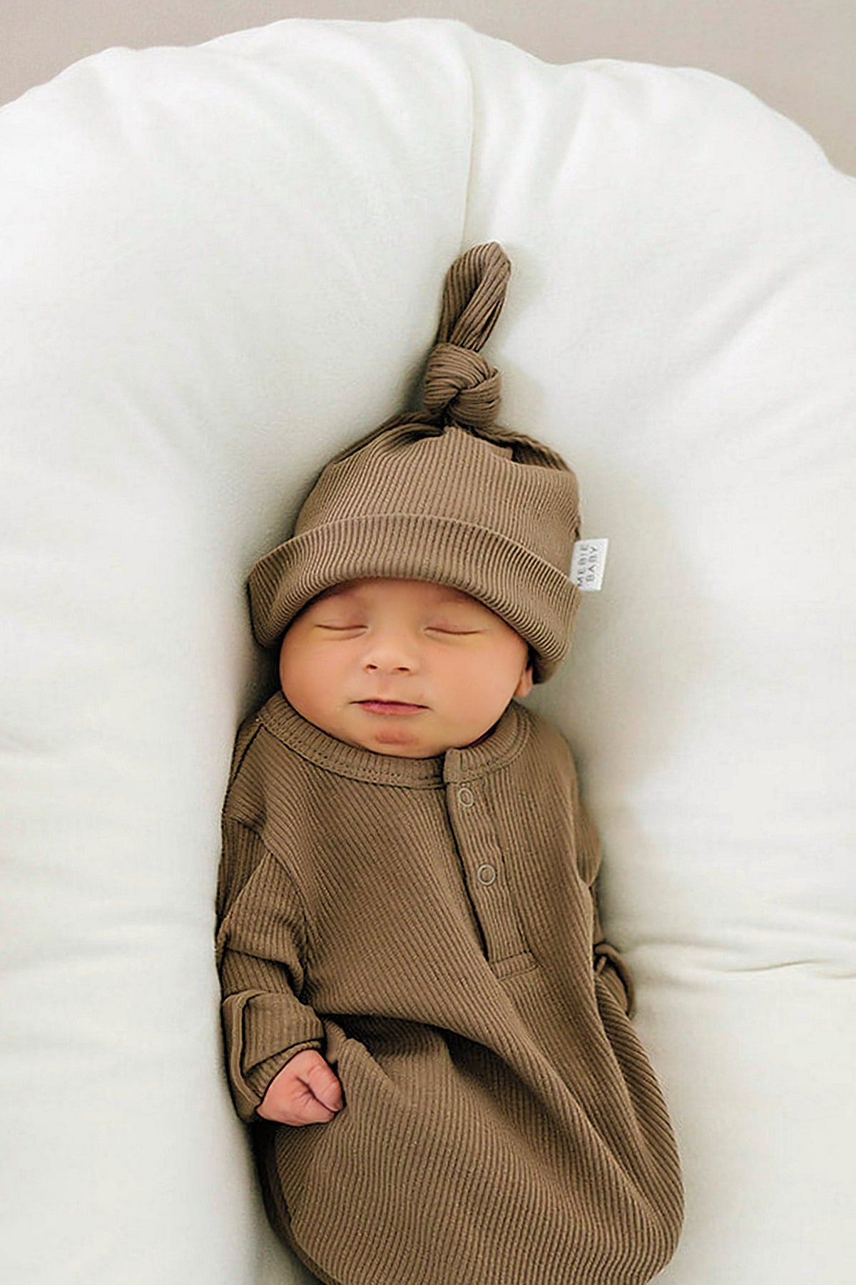 Newborn in a cocoa-colored ribbed outfit with a knotted hat, lying on a white cushion.