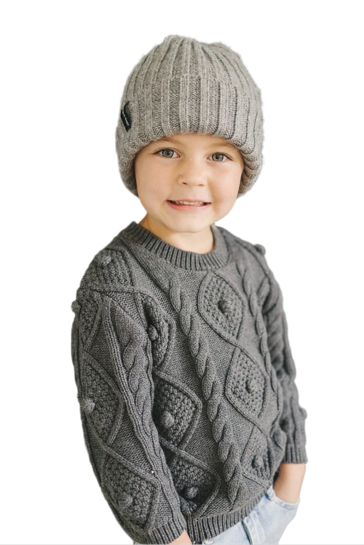 A smiling child wears a grey knit beanie and a patterned grey sweater, showcasing a cozy and stylish winter look.