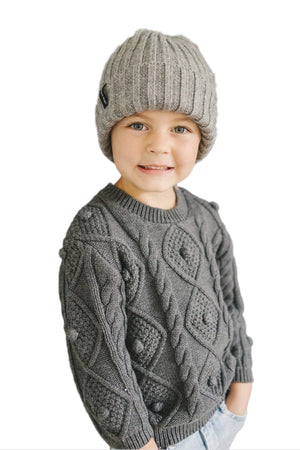 A smiling child wears a grey knit beanie and a patterned grey sweater, showcasing a cozy and stylish winter look.