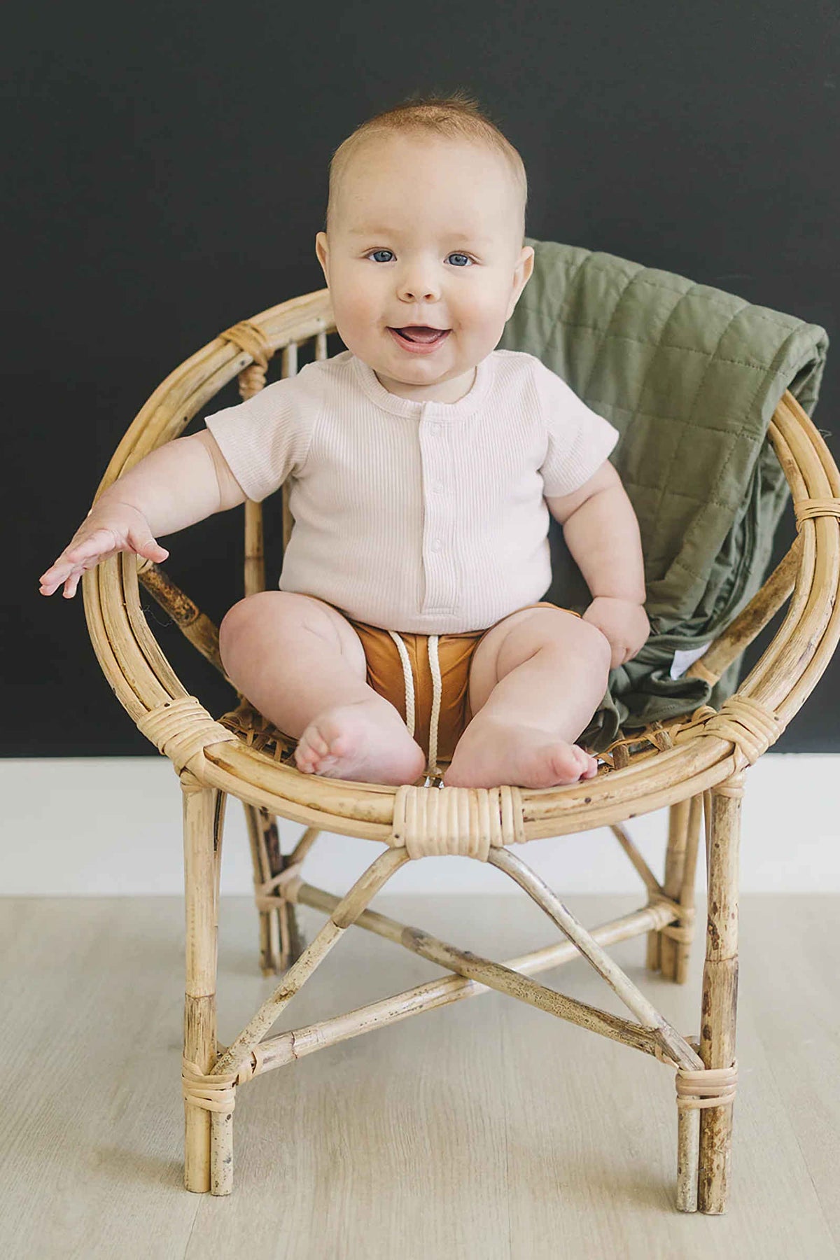 A baby in a ribbed oatmeal bodysuit sits in a wicker chair, with a green quilt draped behind.