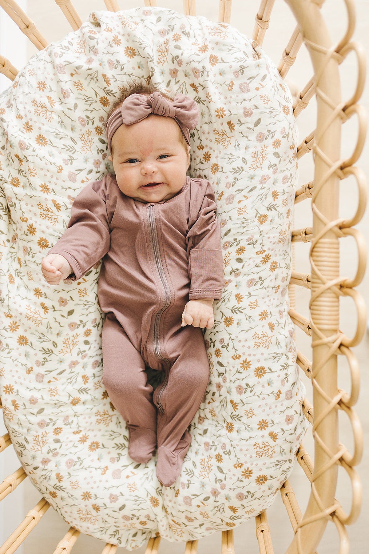 A baby in a plum-colored footed zipper pajama lies in a floral-patterned bassinet.