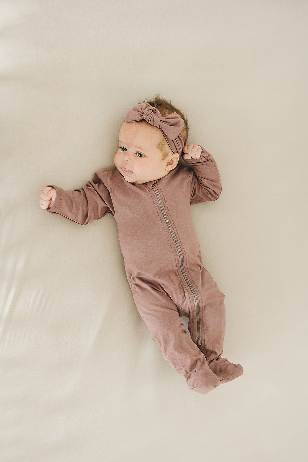 A baby wearing a plum-colored footed zipper pajama with a matching headband lies on a soft beige surface.
